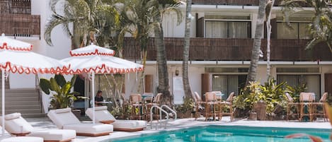 Un área de piscina con sillones de descanso y sombrillas, rodeada de palmeras y un edificio de varios pisos con balcones.
