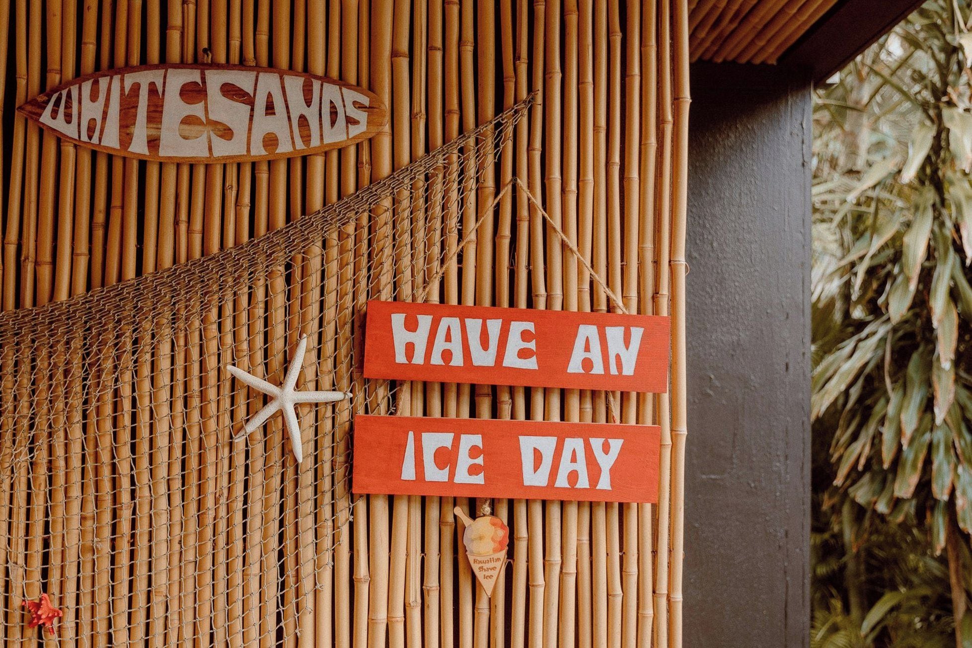 A bamboo wall with a sign that reads 'WHITE SANDS' and two red signs that say 'HAVE AN ICE DAY.'