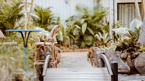 Una terraza de madera que lleva a una casa de paredes blancas, rodeada de plantas tropicales y una pequeña mesa con sillas.