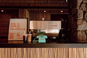 Una cafetera con un panel de menú que muestra tipos de café y precios, un teléfono y una pared de piedra.