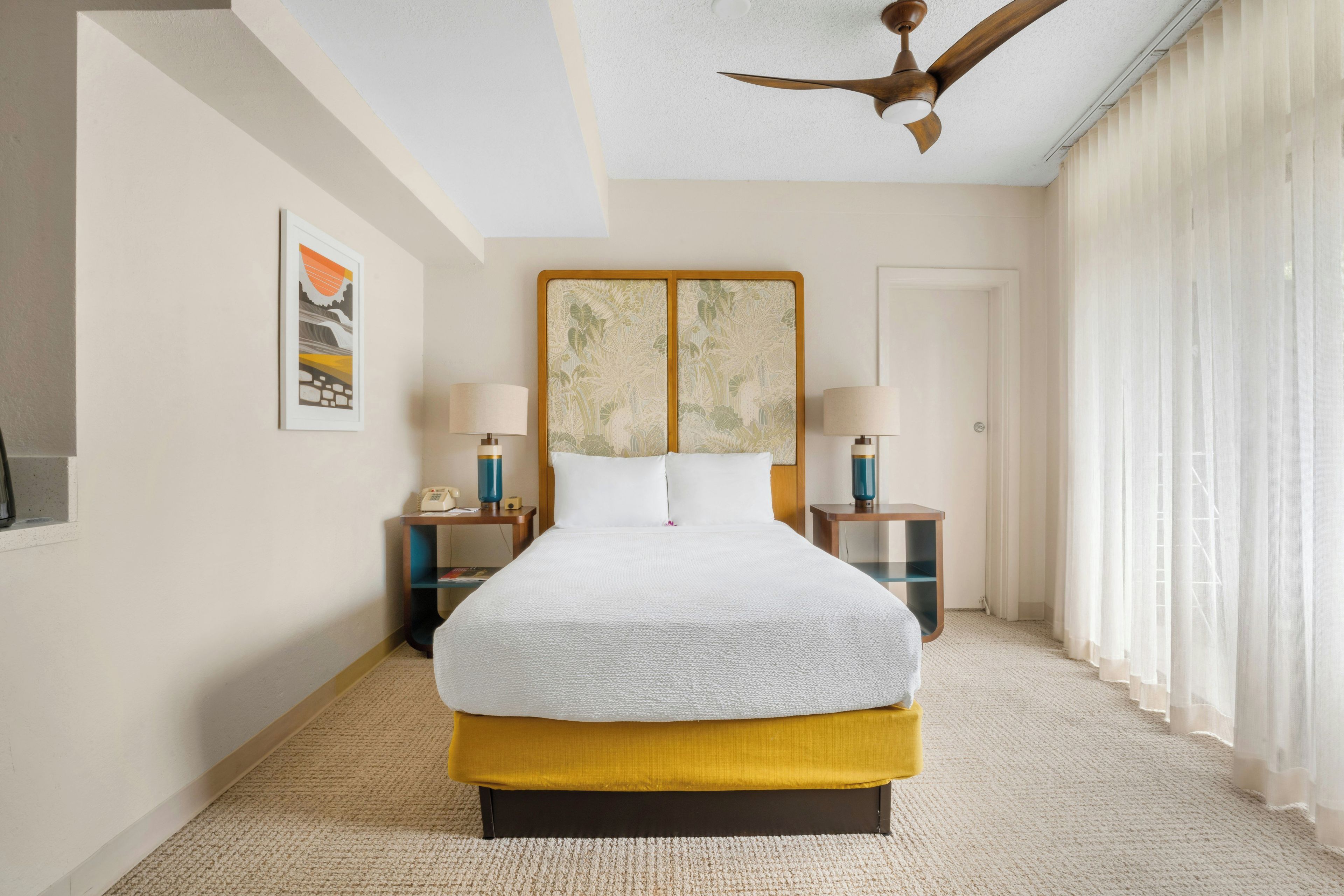 Une chambre d’hôtel avec un grand lit, deux tables de chevet, un ventilateur de plafond et une fenêtre avec des rideaux.