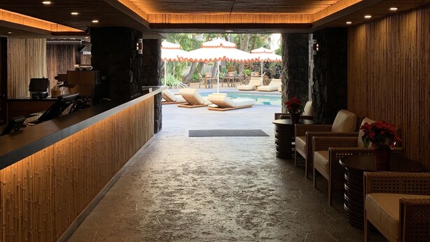 Zona de recepción con mostrador de madera, área de estar con sillas y una mesa, y vista a un área de piscina exterior con sombrillas.