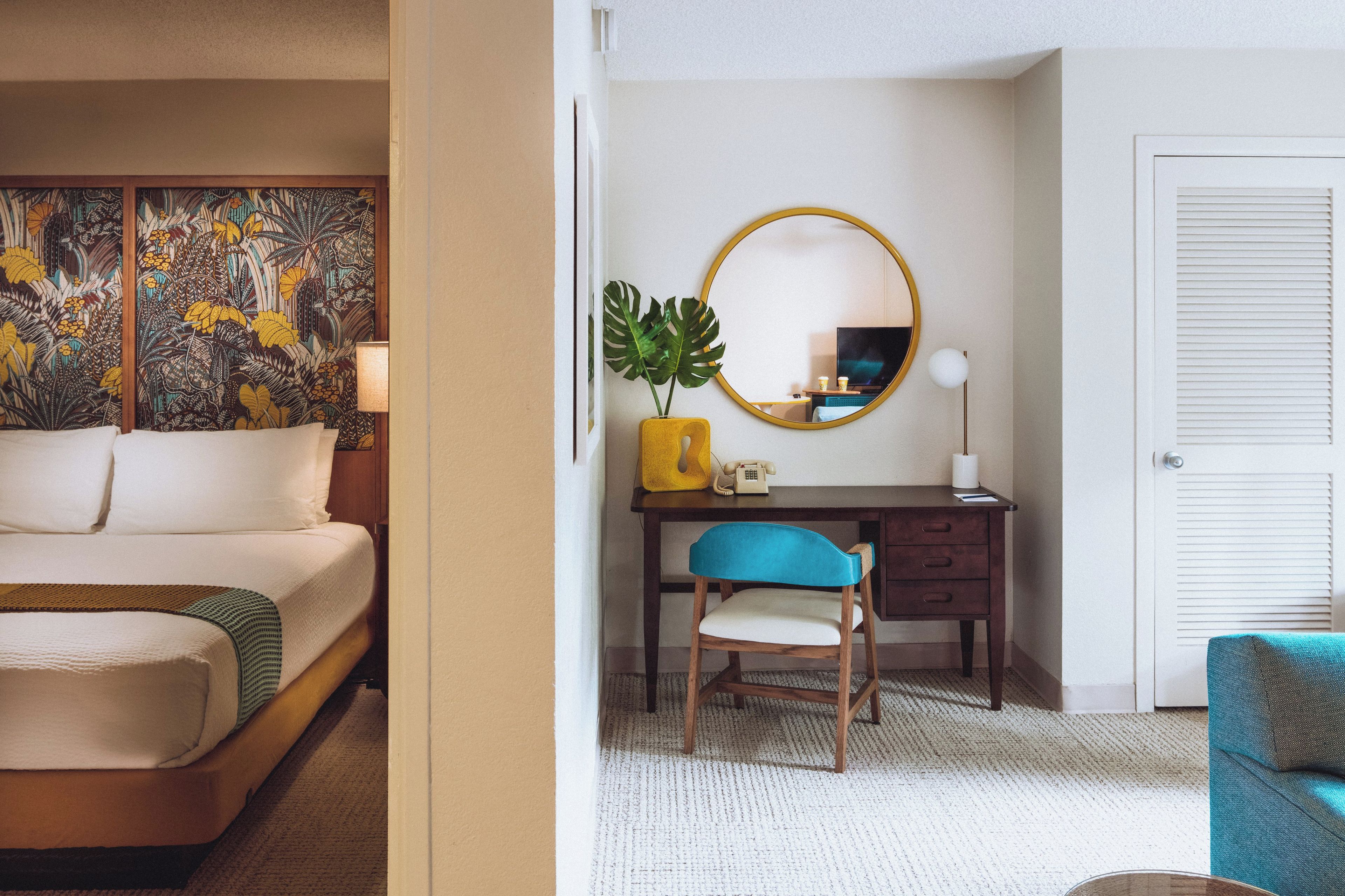 A hotel room with a bed, a desk with a chair, a round mirror, a plant, and a door.