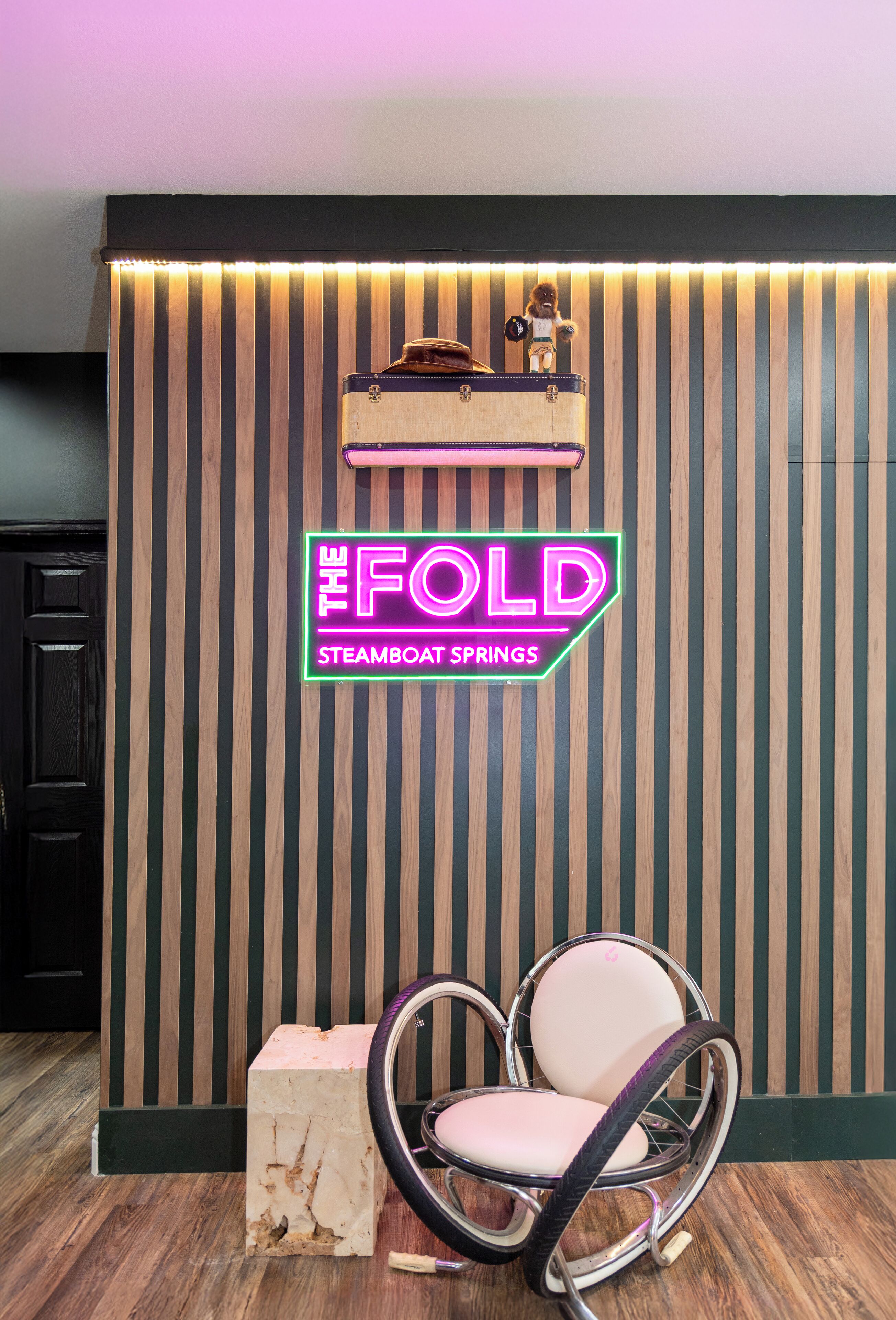 A reception desk with a neon sign reading "The Fold Steamboat Springs" and a vintage suitcase on top.