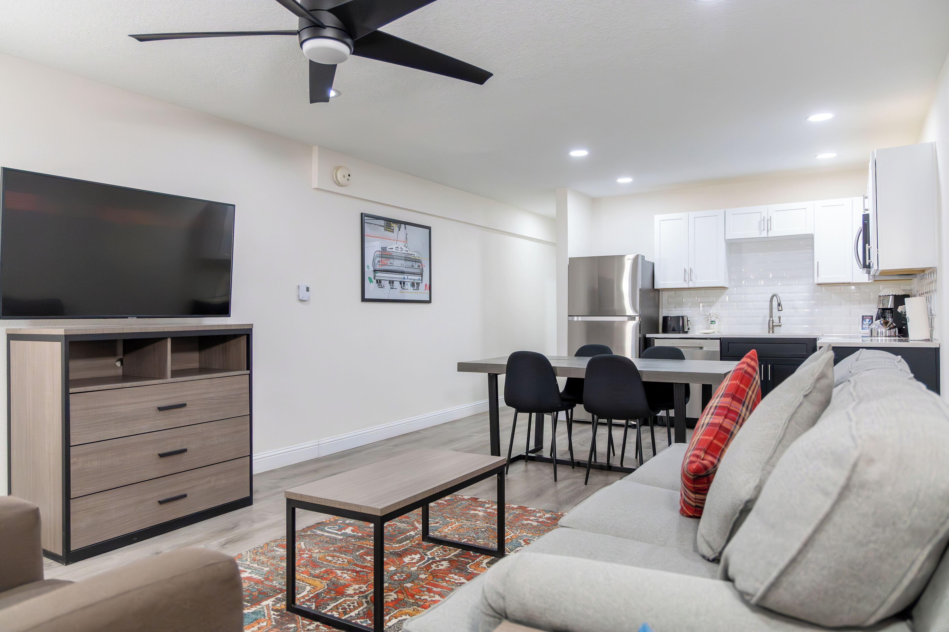 A modern living room with a sofa, a coffee table, and a TV. There is a dining area with chairs and a table. The kitchen has white cabinets and a stainless steel range hood.