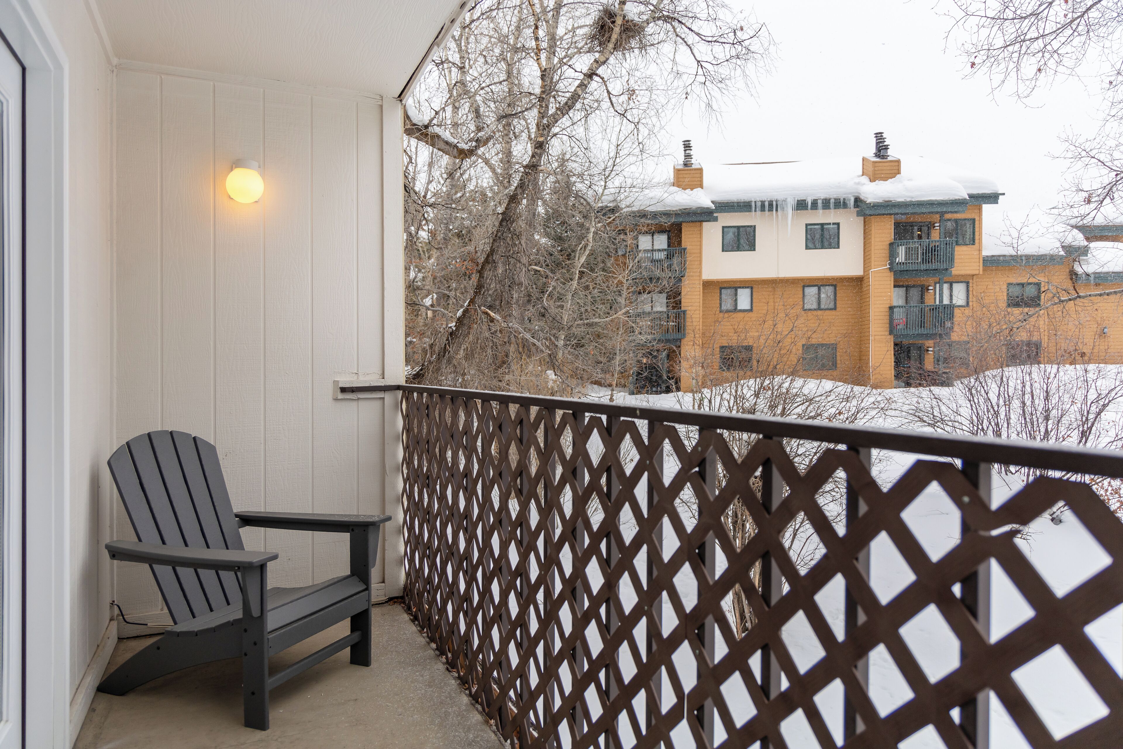 Un balcón con baranda de celosía de madera, una silla Adirondack y vistas de edificios con nieve en los tejados.