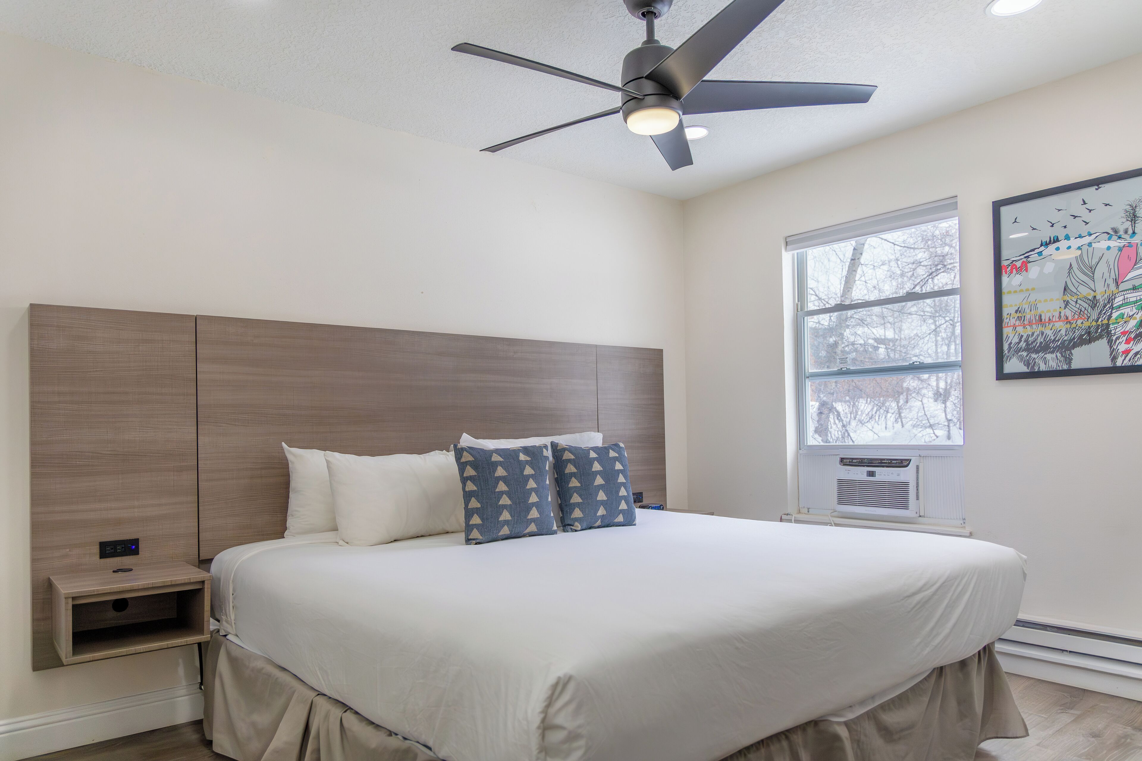 A bedroom with a large bed, a ceiling fan, a nightstand, and a window with a view of trees.