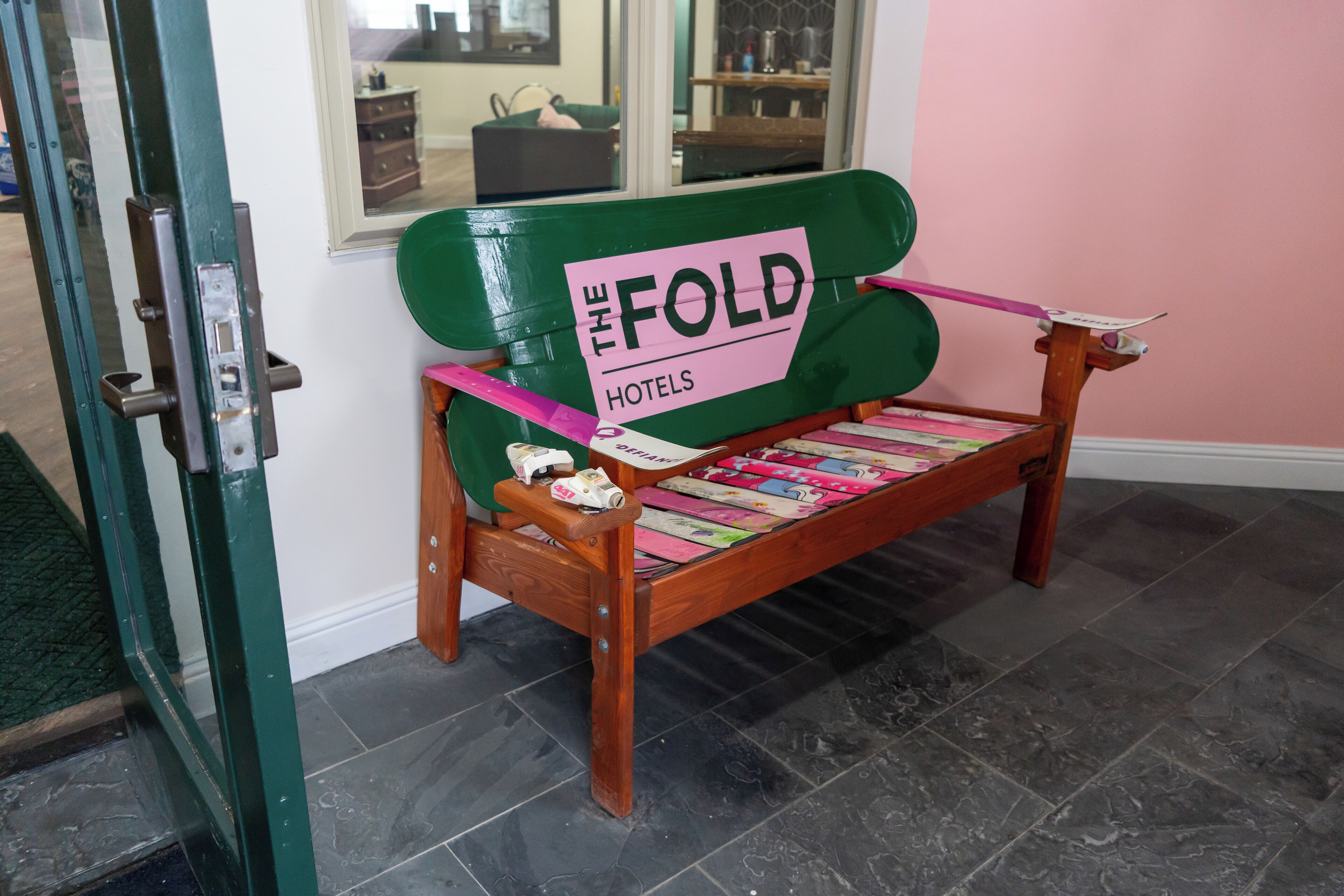 A green bench with a sign reading "The Fold Hotels" and a magazine rack attached to it.