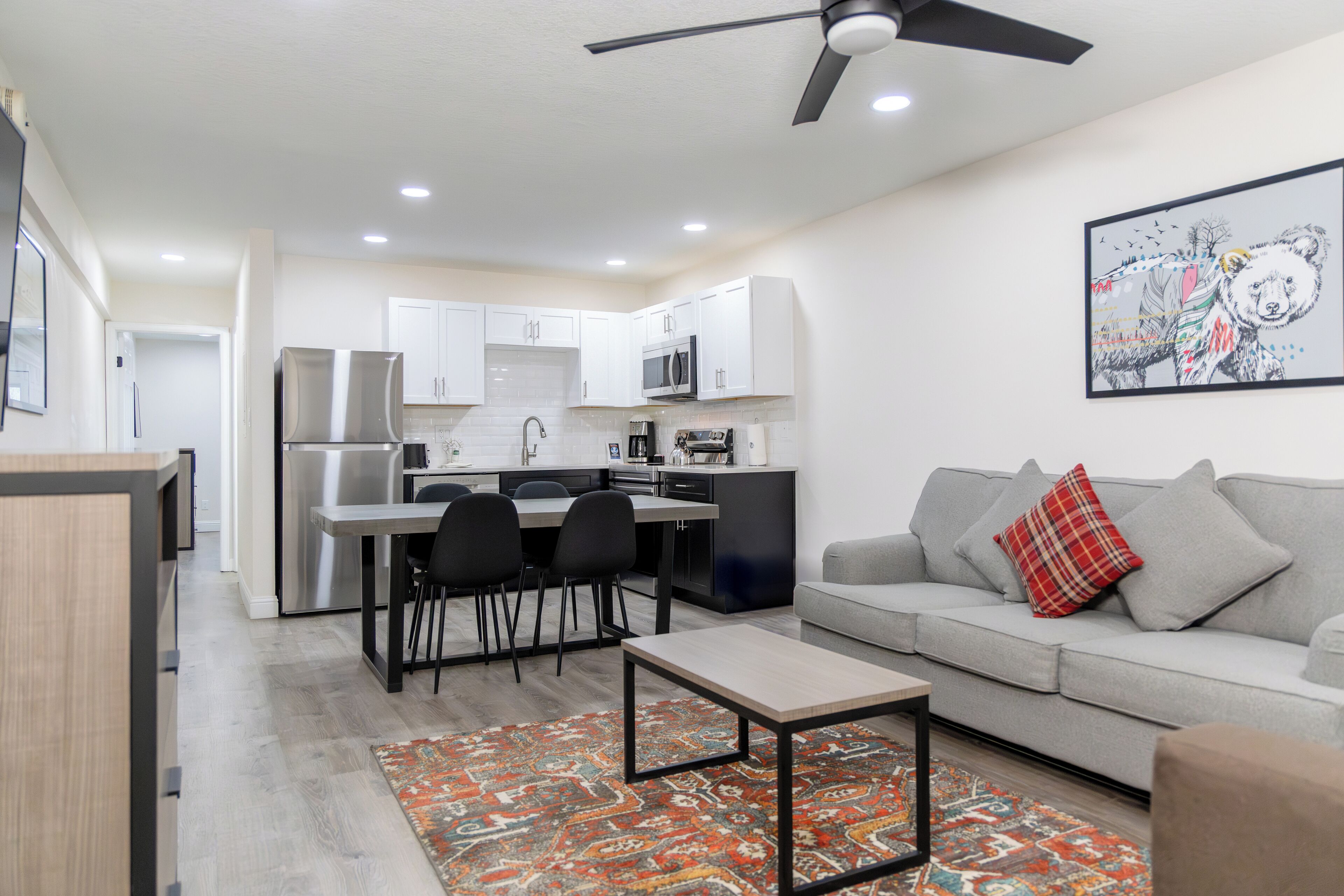 A modern kitchen with white cabinets, a dining area with black chairs, and a grey sofa with a red pillow.