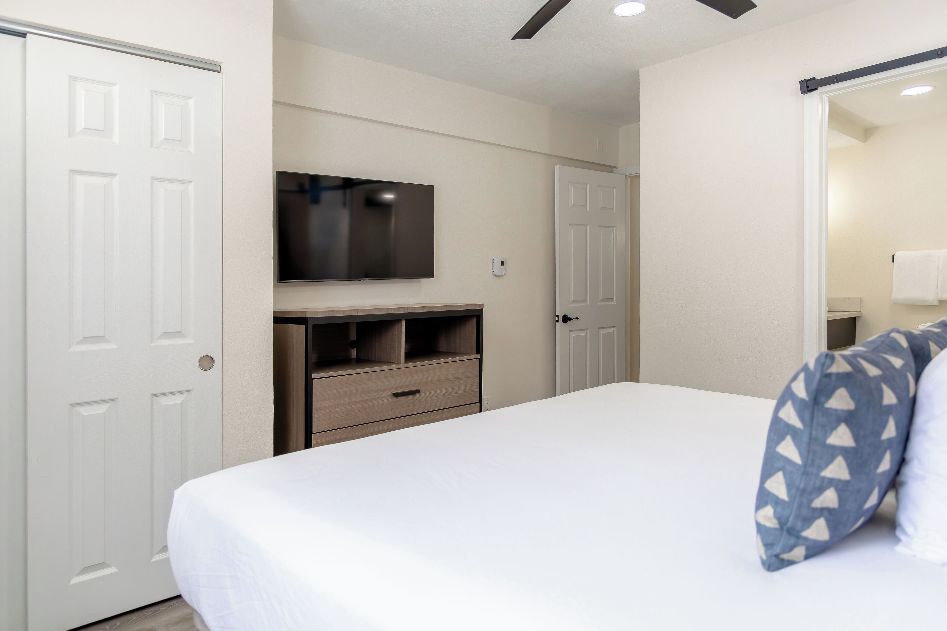 Un dormitorio con cama, televisor montado en la pared, armario y puerta que da al baño.