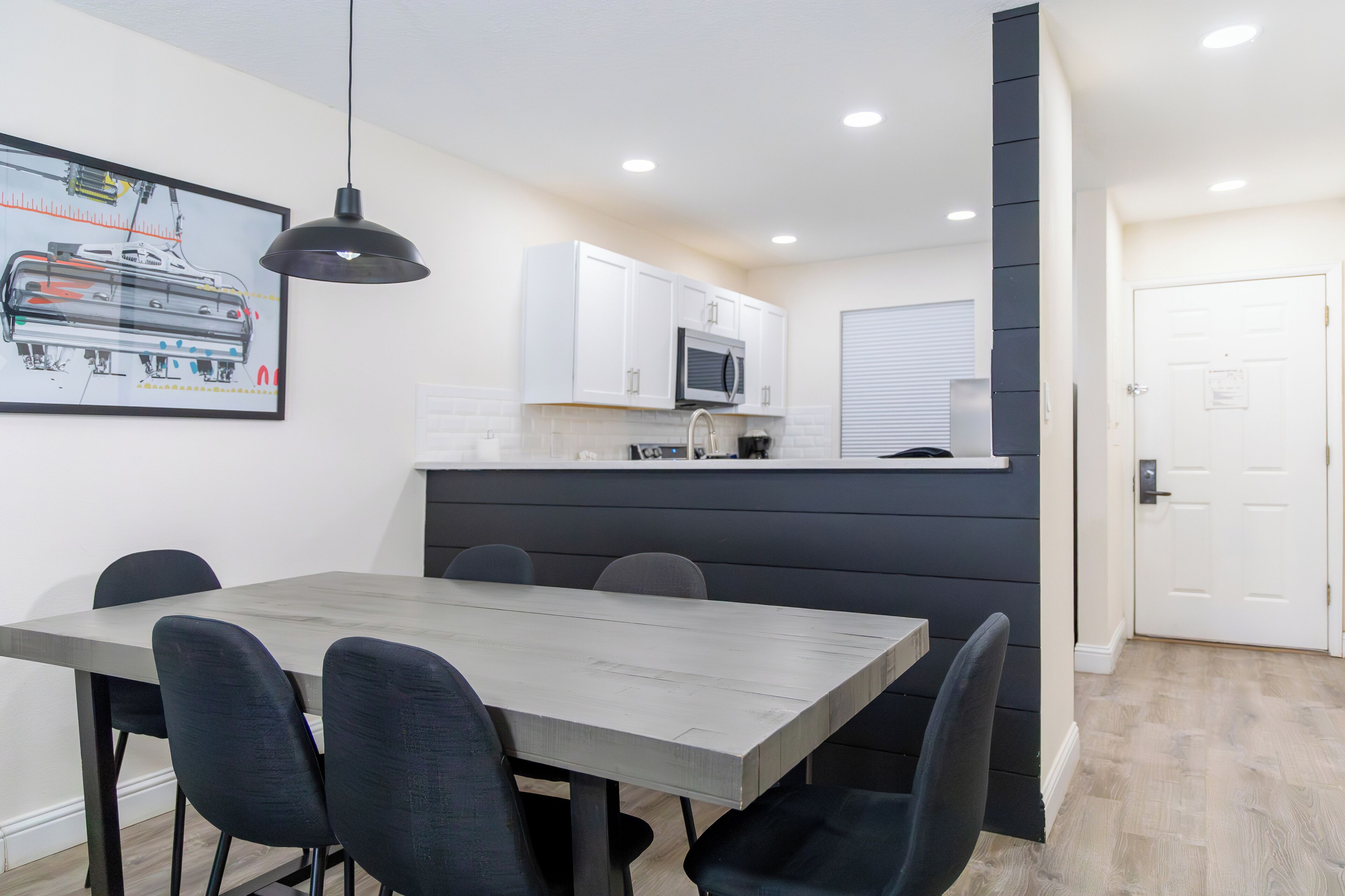 Una cocina moderna con armarios blancos, un microondas y un lavamanos. Hay un comedor con una mesa de madera y sillas negras.