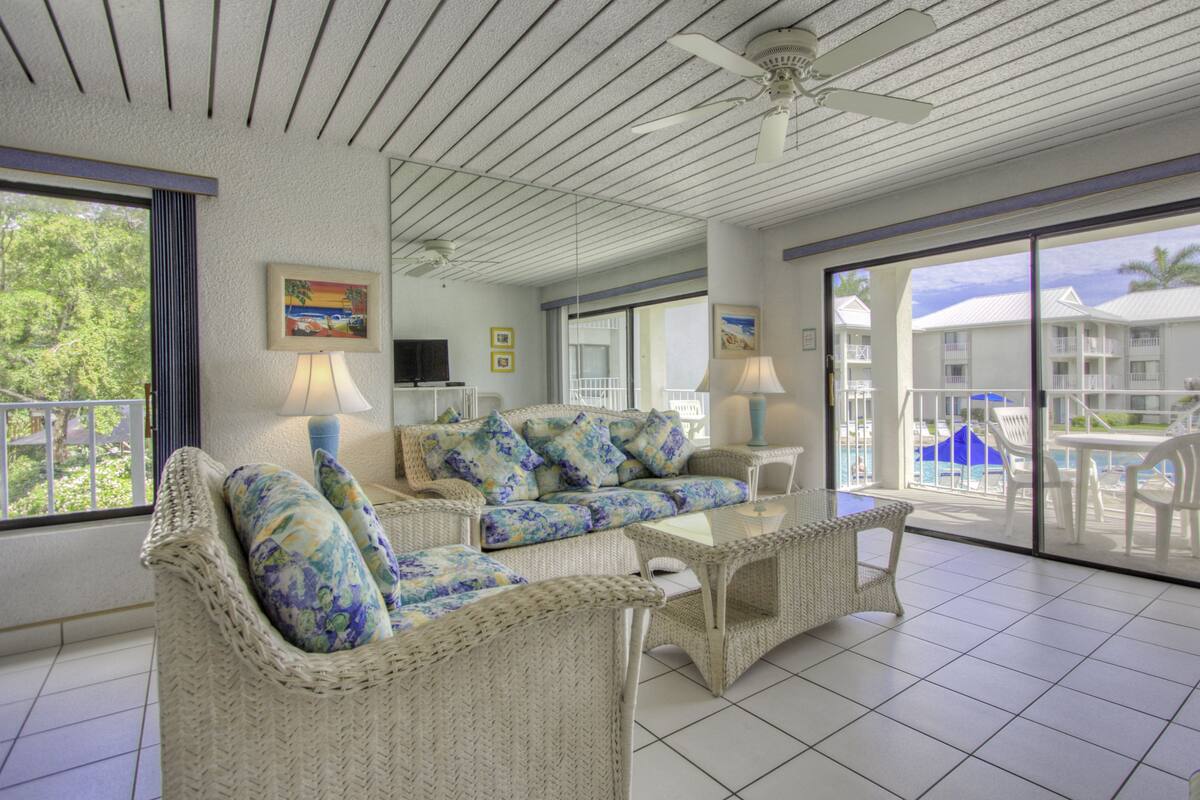 A spacious balcony with wicker furniture, a ceiling fan, and a view of a pool and buildings.