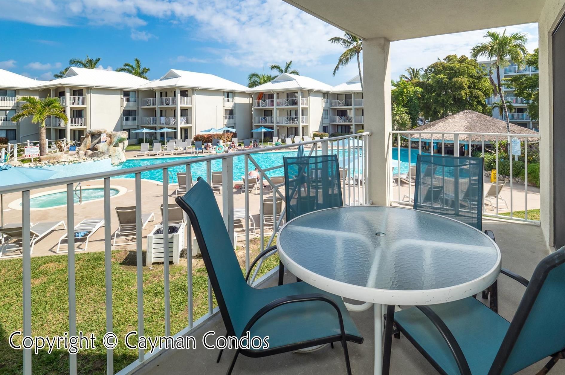 A balcony with a glass table and chairs overlooking a pool area with lounge chairs and umbrellas.