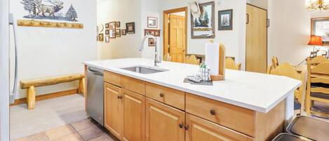 A kitchen with wooden cabinets, a white countertop, and a sink. There is a bench and a door leading to another room.