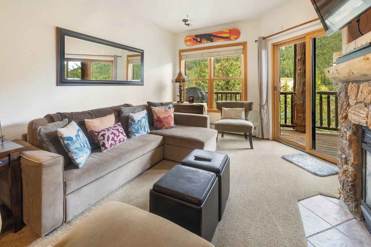 A living room with a sectional sofa, ottomans, a fireplace, and a large window with a view of greenery.
