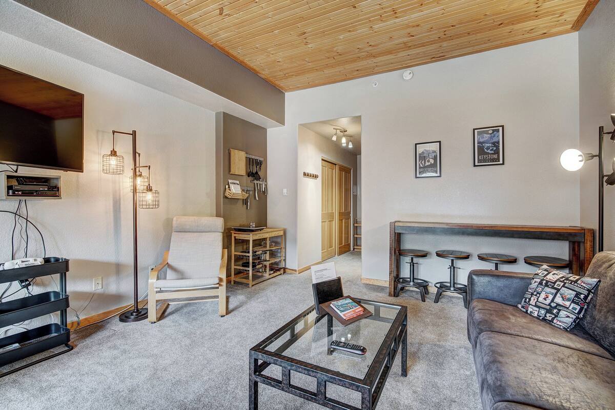 A modern living room with a flat-screen TV, a glass coffee table, a sofa, a chair, and a bar area with stools.