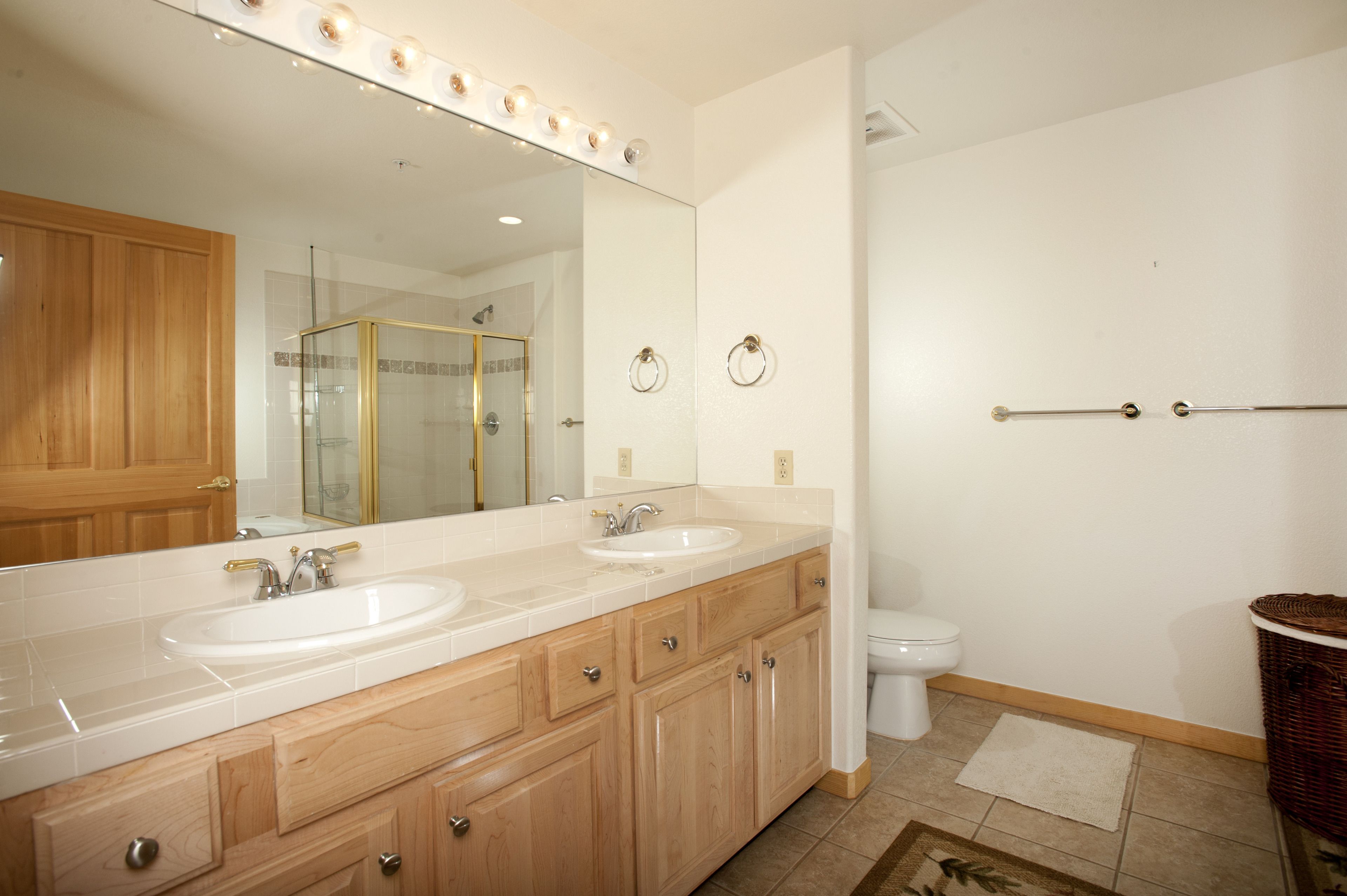 A bathroom with two sinks, a large mirror with lights, a shower, and a toilet.