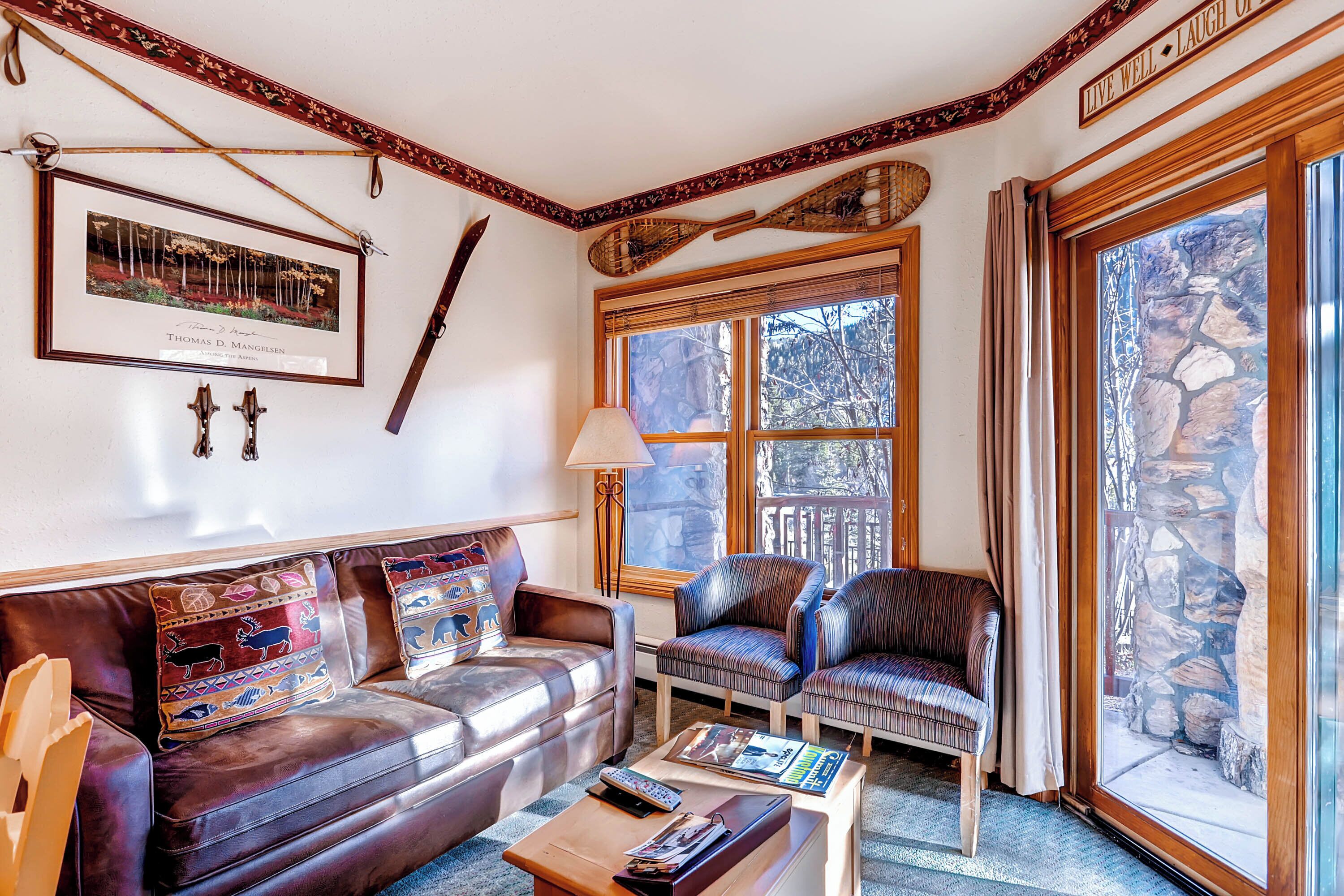 A cozy living room with a leather couch, patterned pillows, a wooden coffee table, and a large window with a stone wall view.