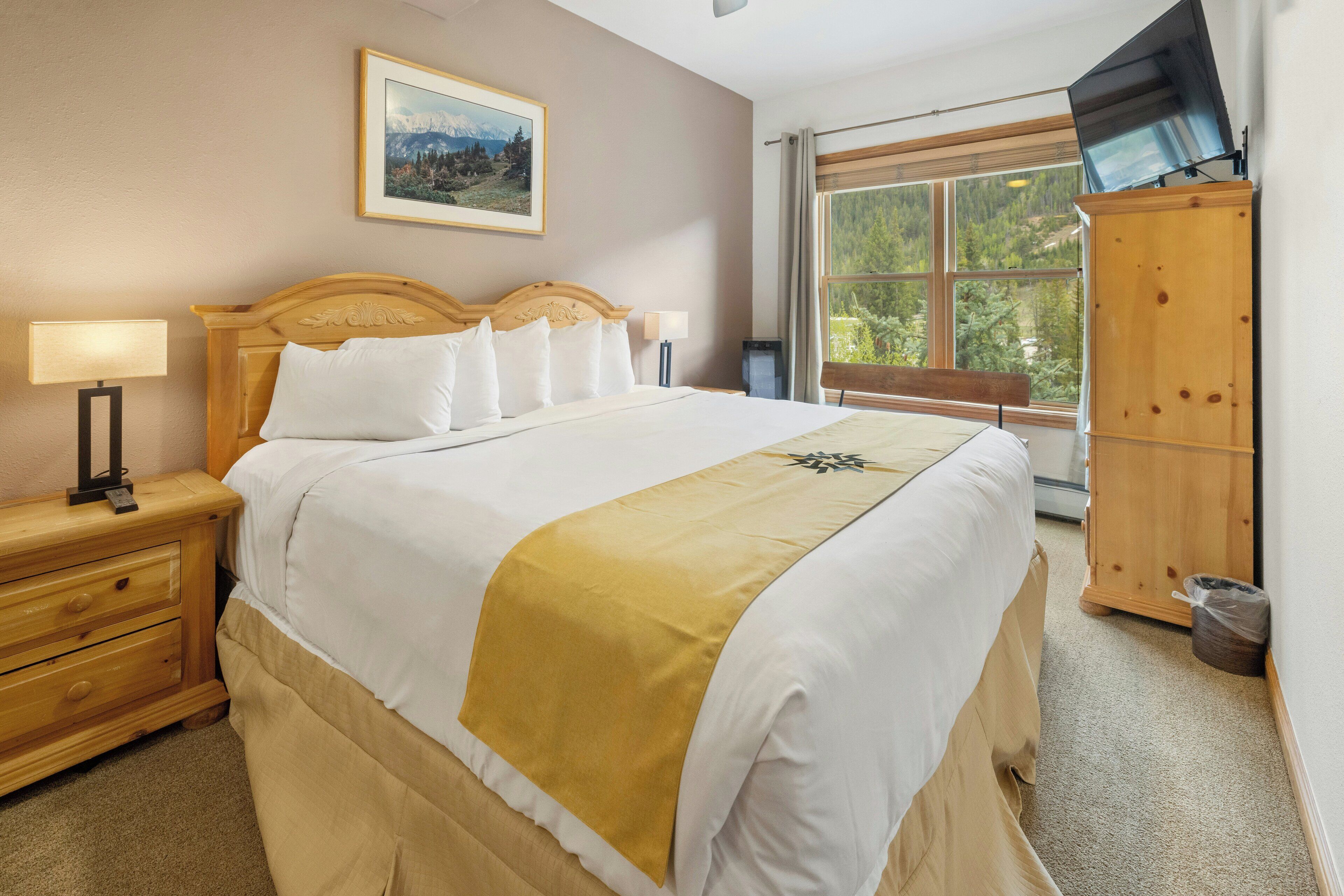 A neatly made bed with a white and yellow bedspread, a wooden nightstand with a lamp, a framed picture on the wall, and a window with a view of trees.