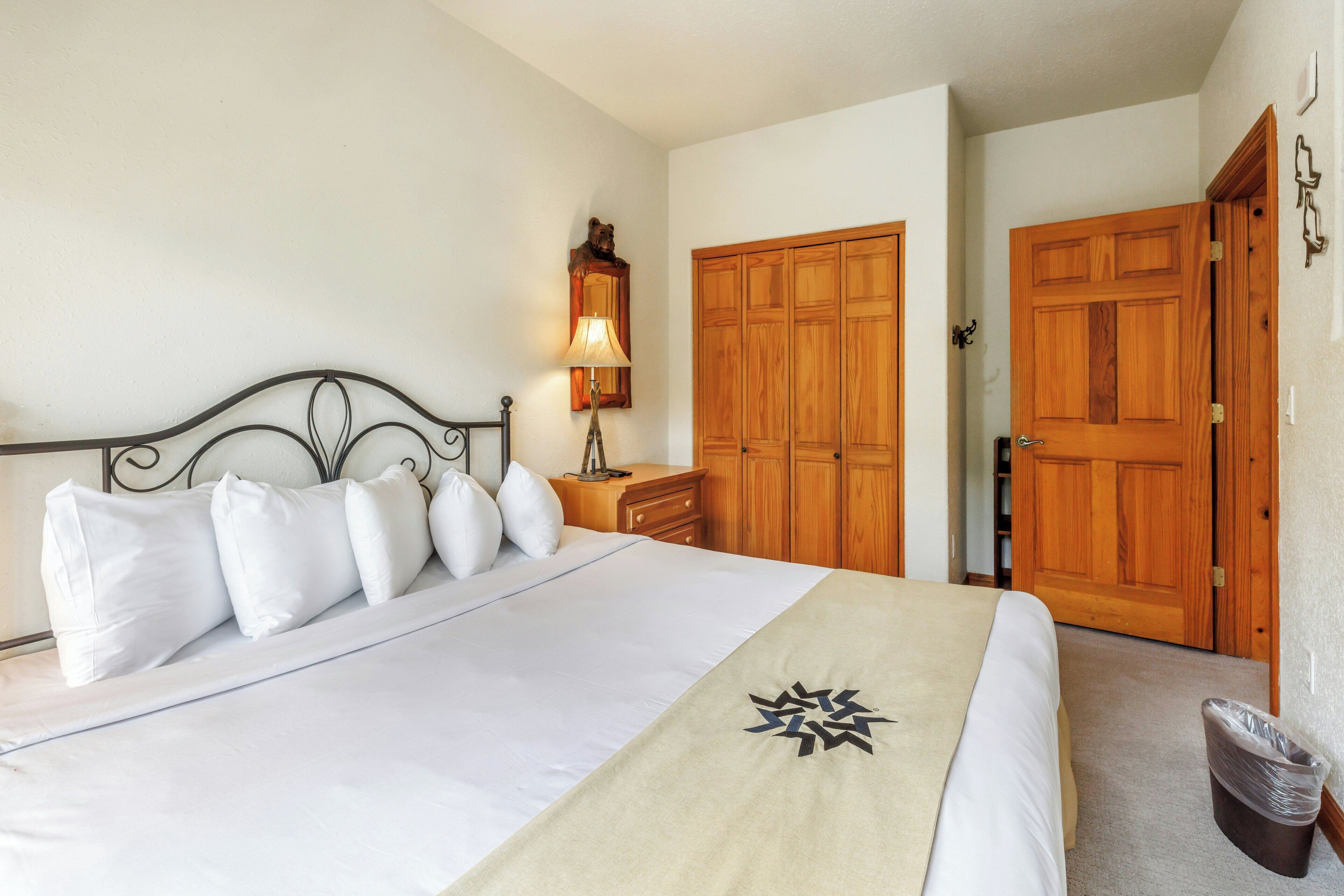A neatly made bed with white linens and a beige throw, a wooden nightstand with a lamp, and a wooden wardrobe.