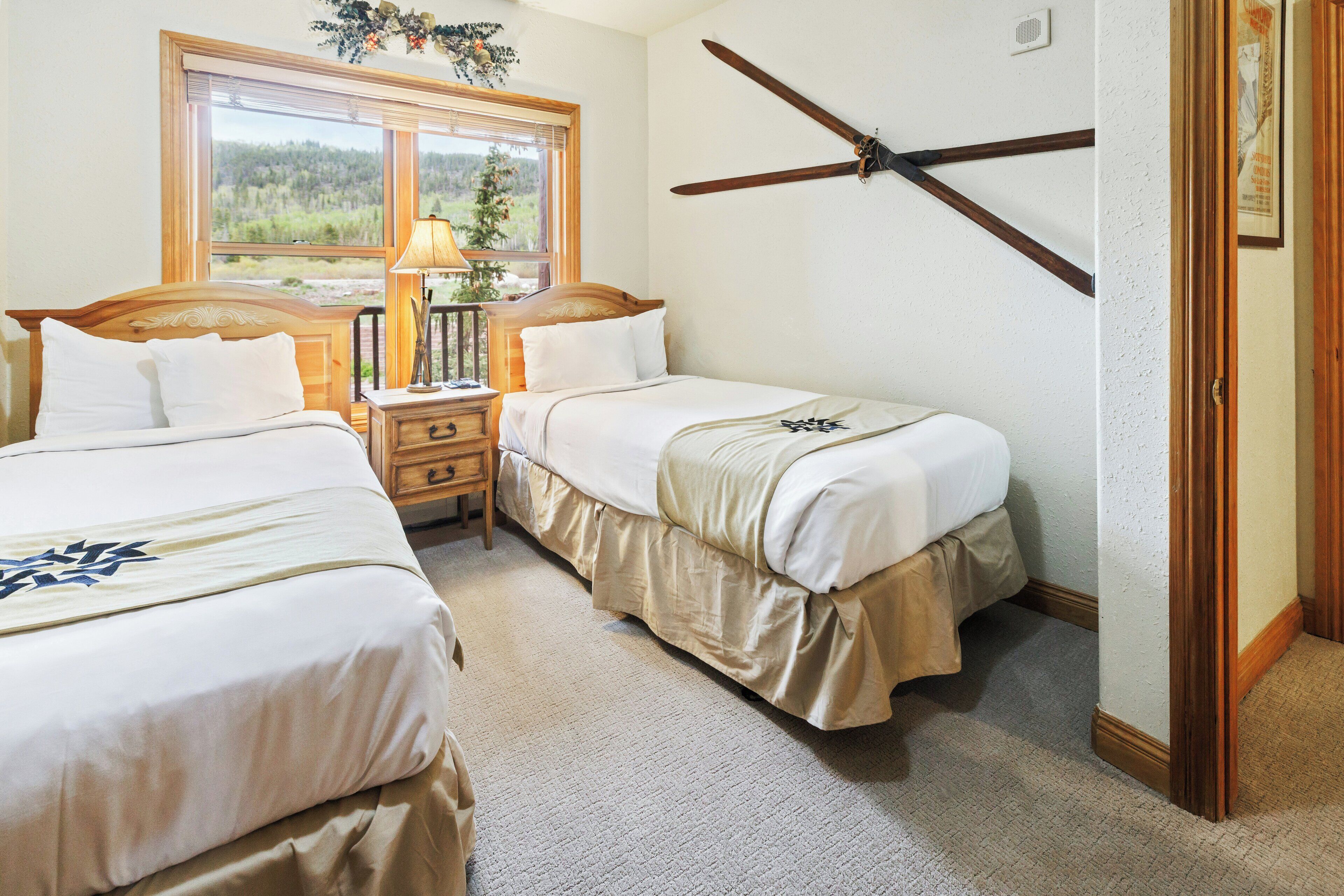 A bedroom with two beds, a nightstand, a window with a view of a forest, and a ceiling fan.