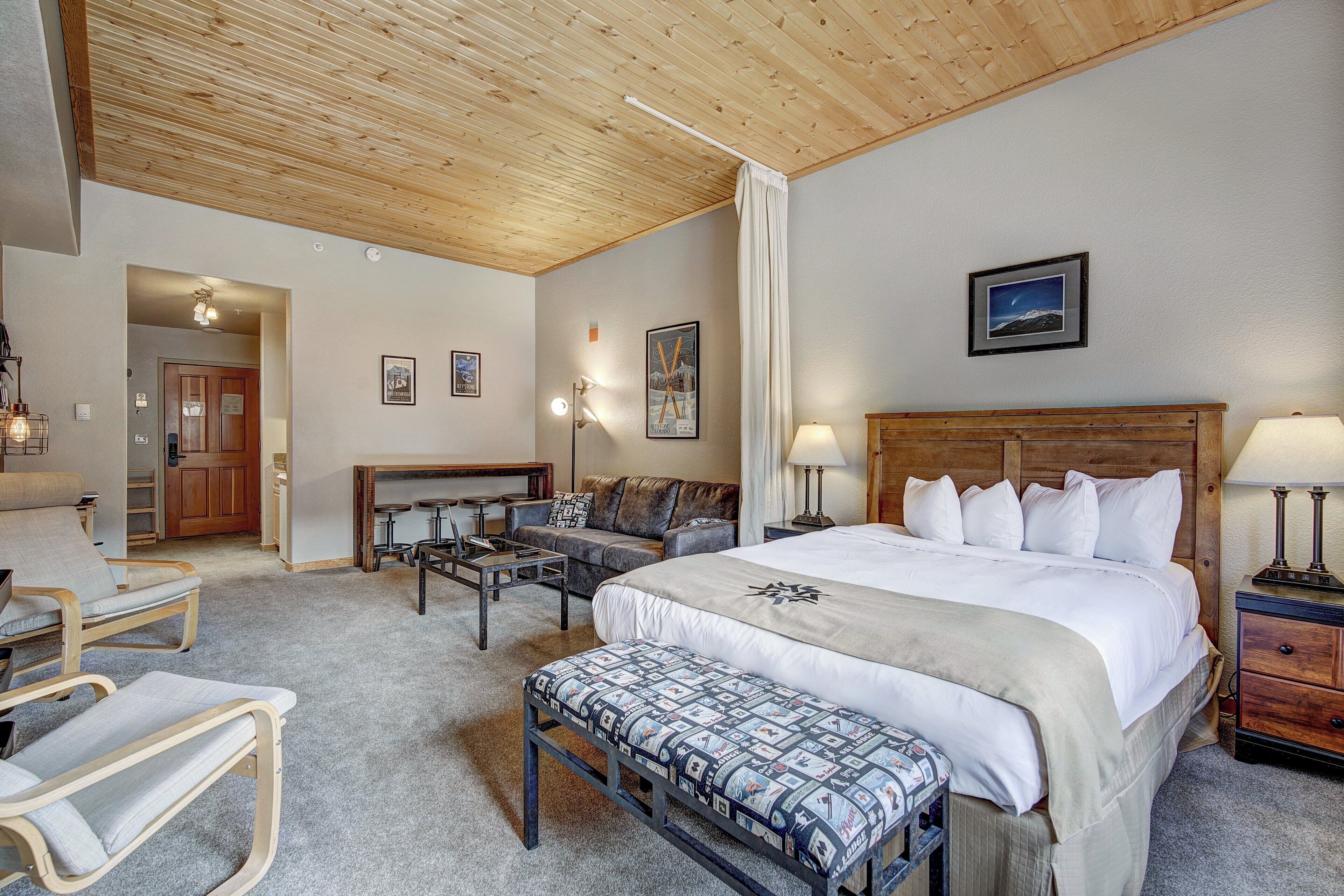 A bedroom with a large bed, a wooden headboard, a bedside table with lamps, a bench, a sofa, and a desk with chairs.
