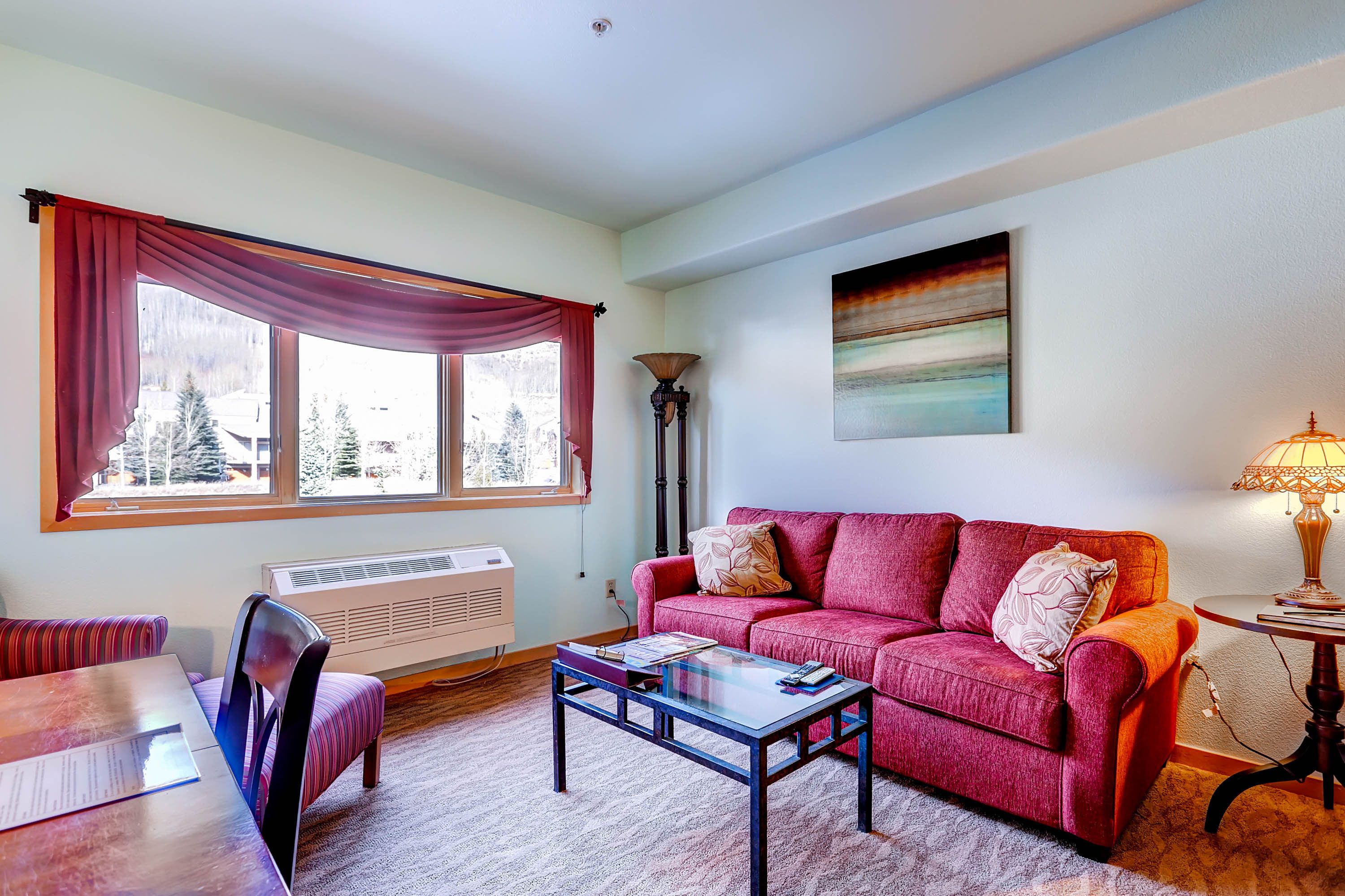 A living room with a red sofa, a glass coffee table, a dining table with chairs, a radiator, and a window with curtains.