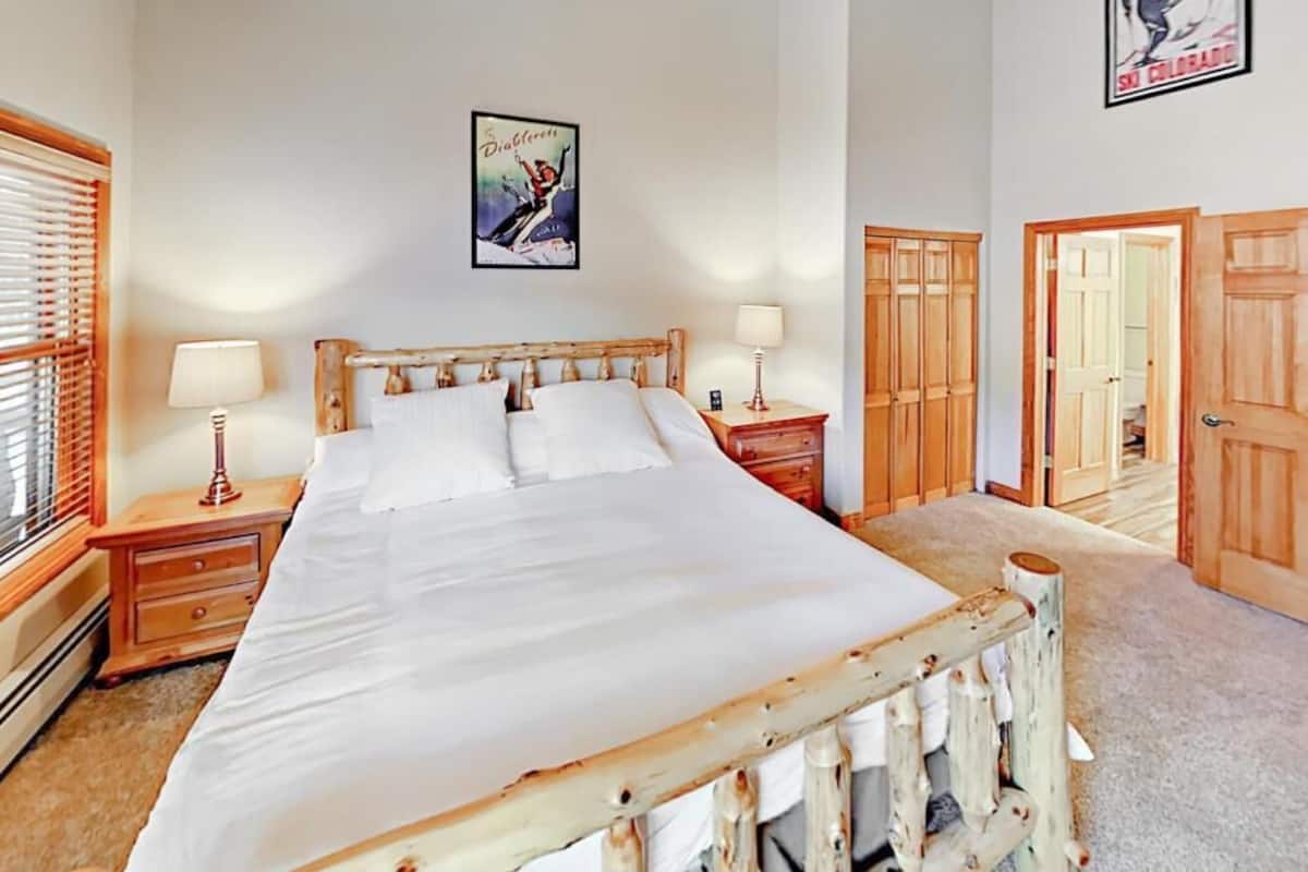 A bedroom with a wooden bed frame, two bedside tables with lamps, a poster on the wall, and wooden doors.