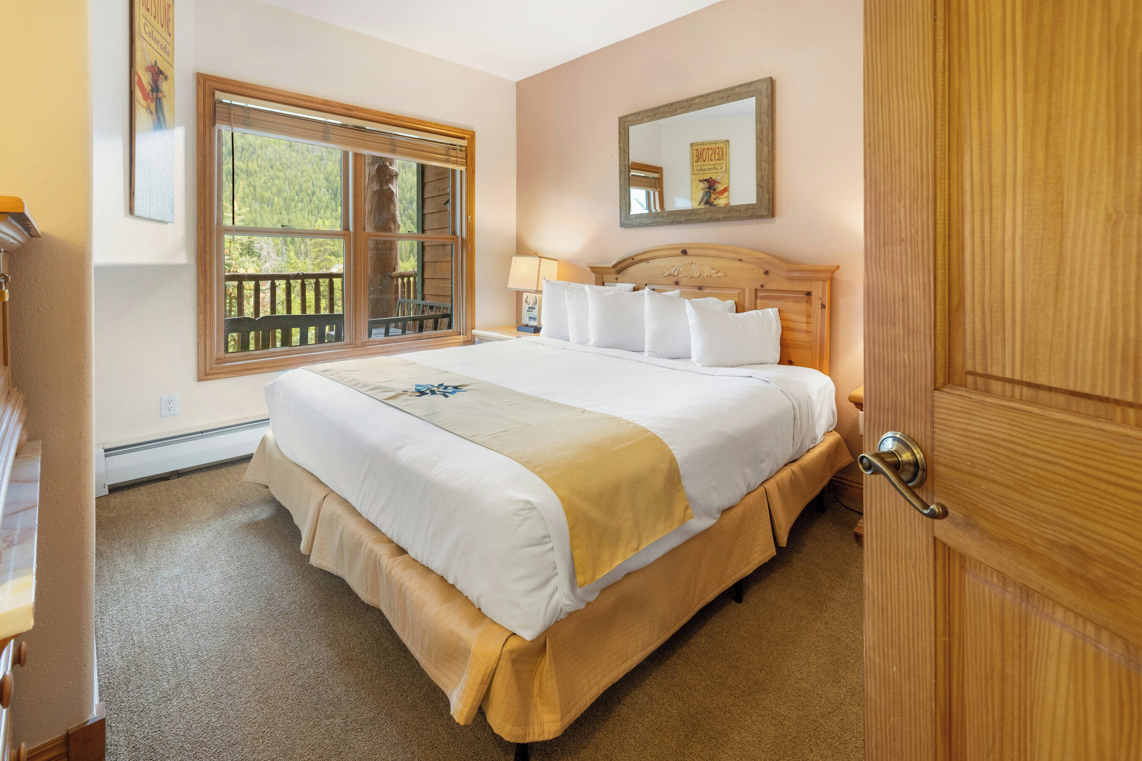 A neatly made bed with white and yellow linens, a wooden headboard, and a window with a view of greenery.