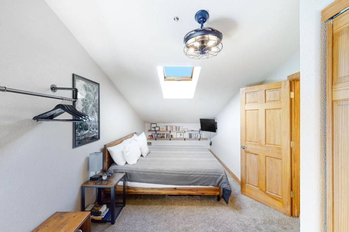 A bedroom with a bed, a desk, a chair, a wardrobe, a ceiling fan, a skylight, and a bookshelf.