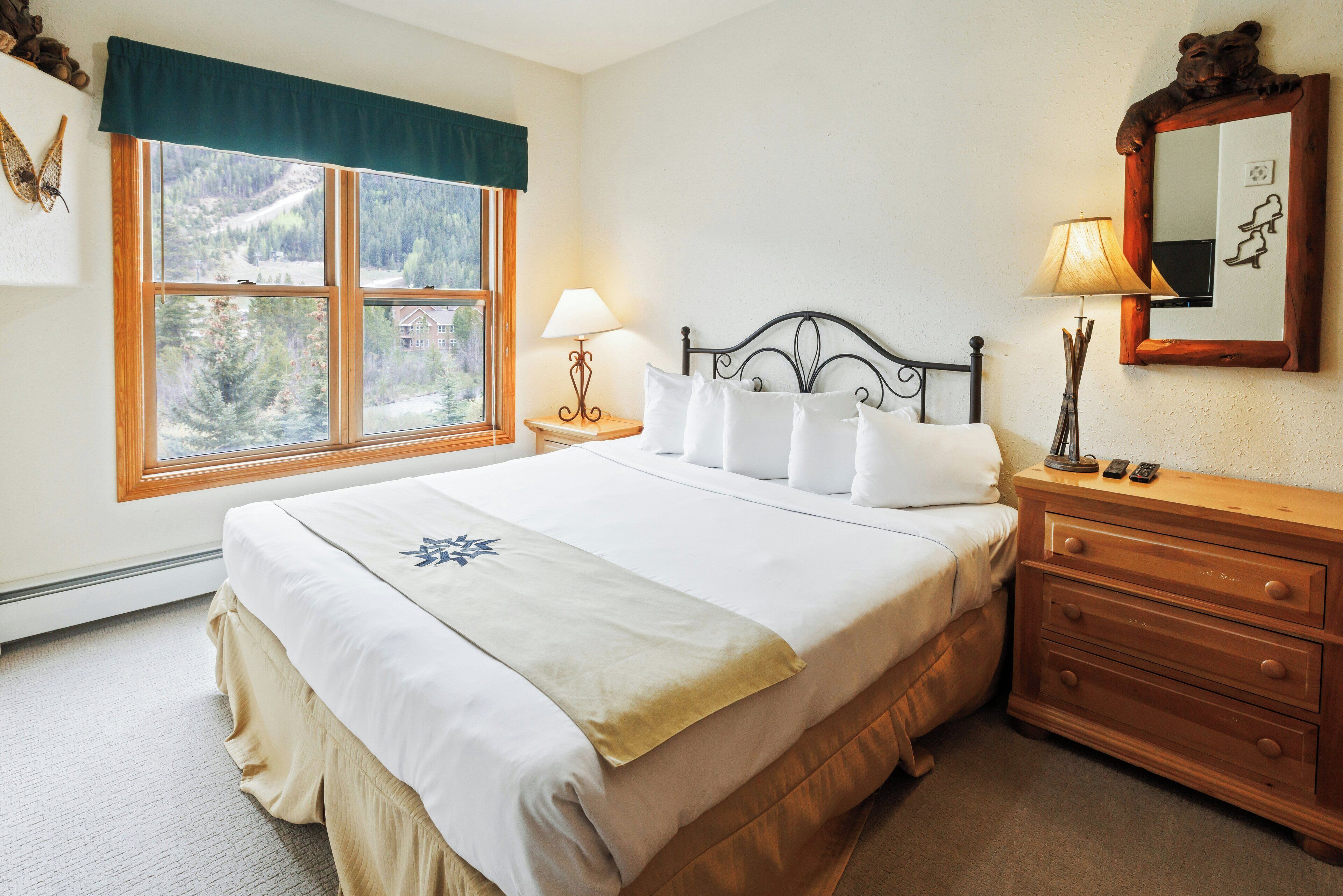 A neatly made bed with white linens and a decorative pillow, a wooden nightstand with a lamp, a dresser with drawers, and a large window with a view of trees and a house.