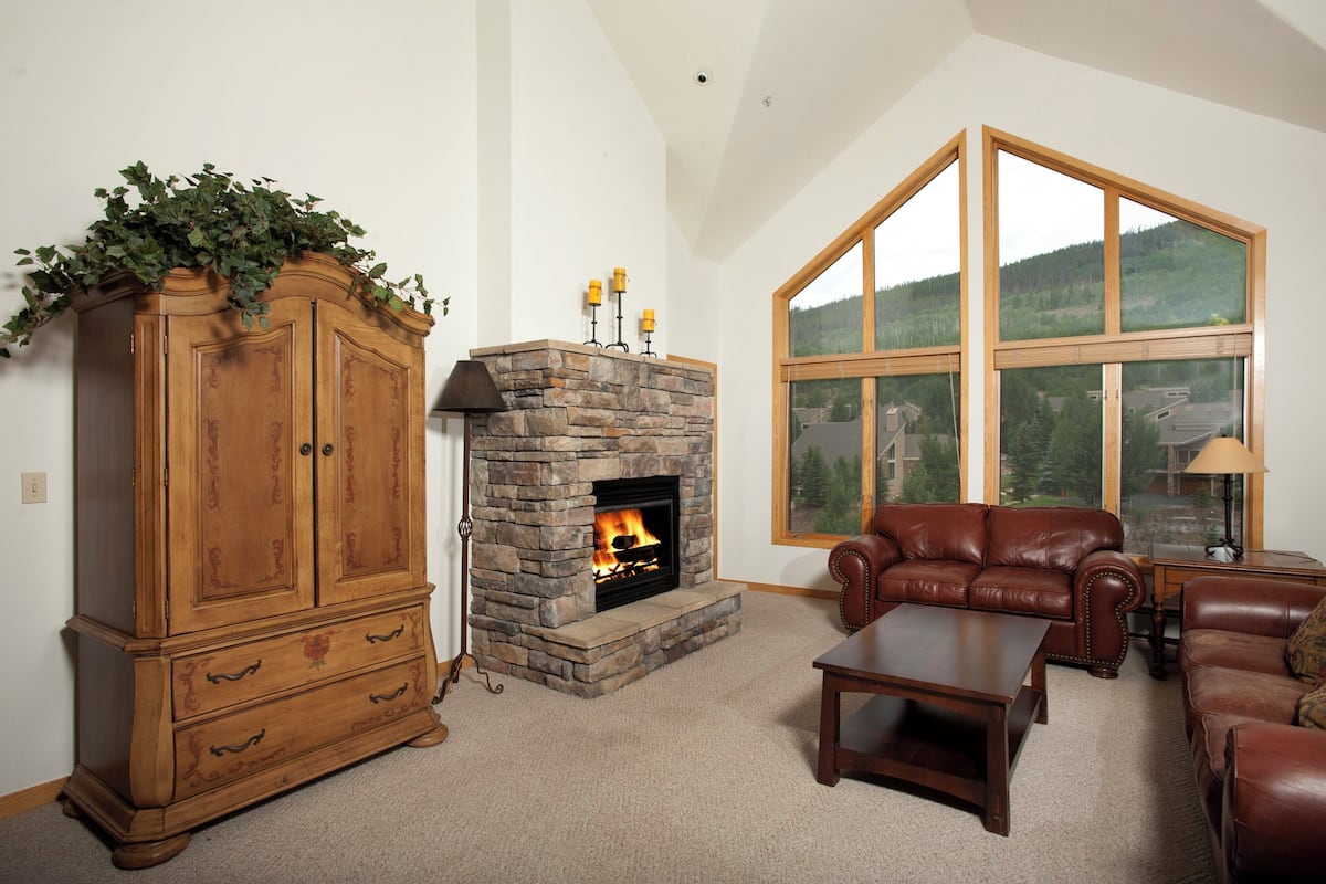 A living room with a stone fireplace, leather furniture, and large windows offering a view of a forested area.