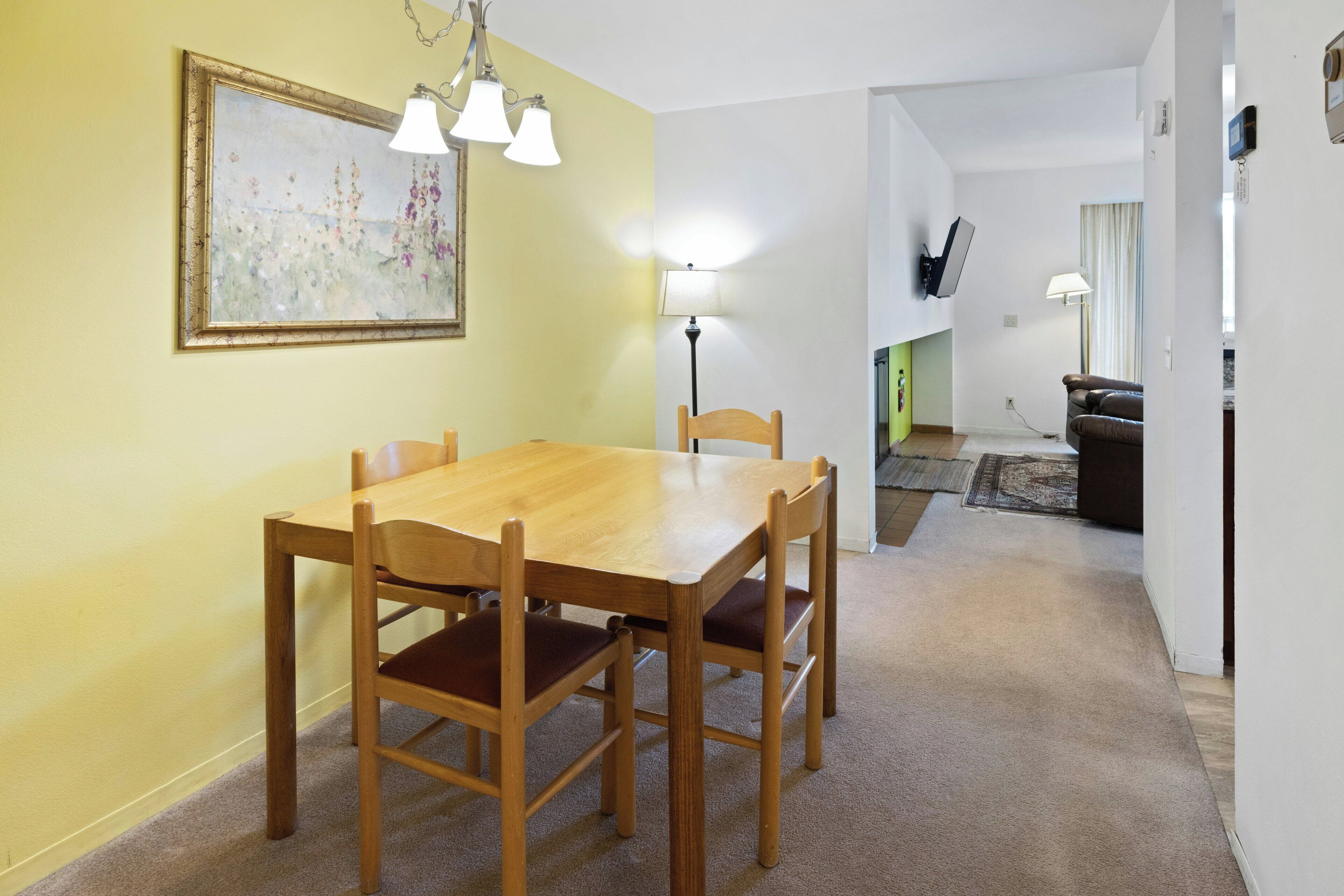 A dining area with a wooden table and chairs, a painting on the wall, and a lamp.