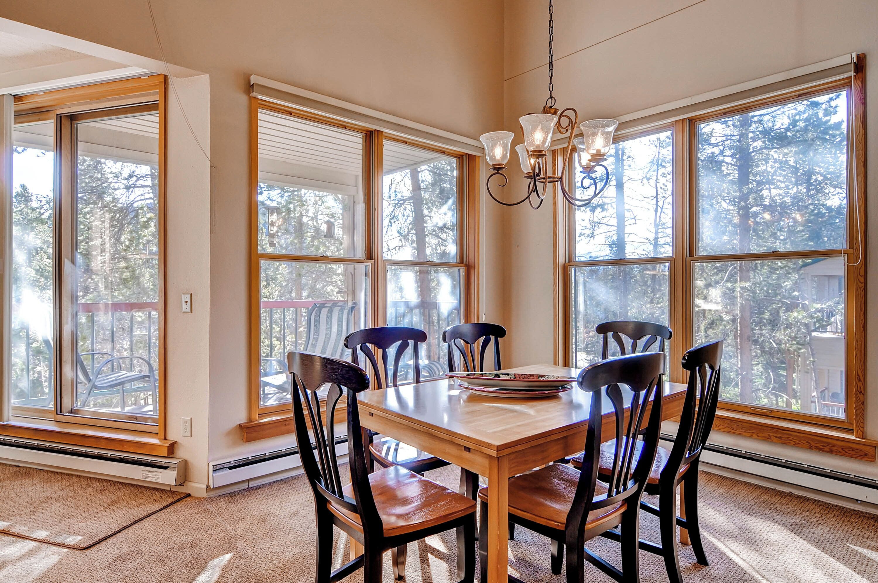 A dining area with a wooden table and chairs, large windows, and a chandelier.