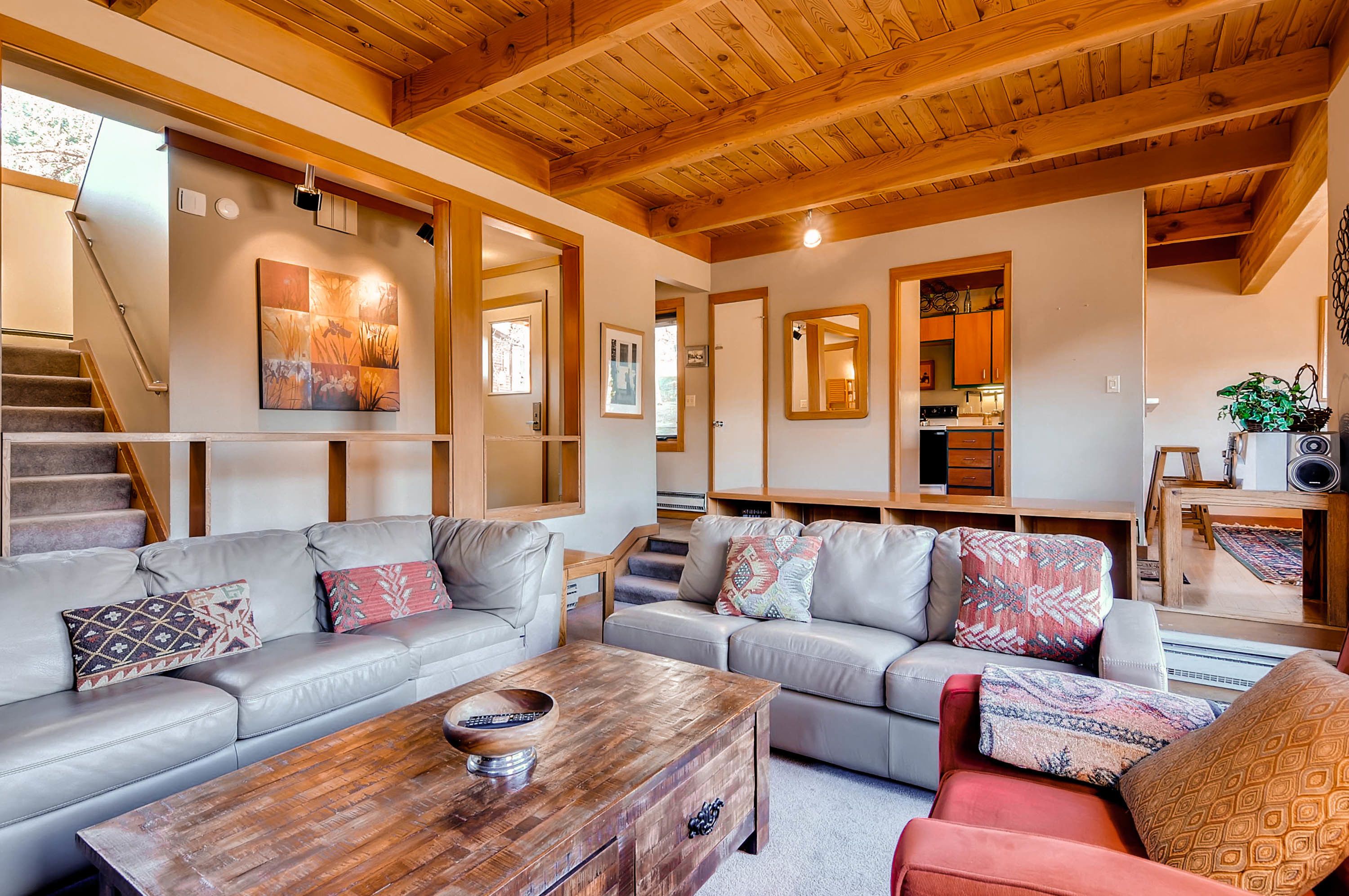 A spacious living area with a wooden ceiling, a staircase, and a kitchen visible through an open door.