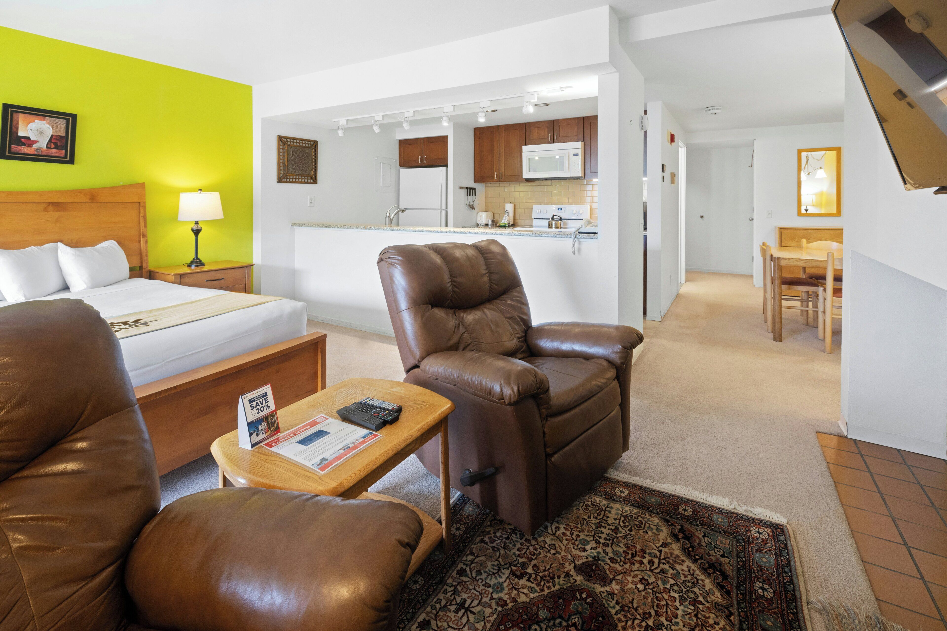 A hotel room with a bed, a brown leather chair, a small table with a remote control, and a kitchen area in the background.