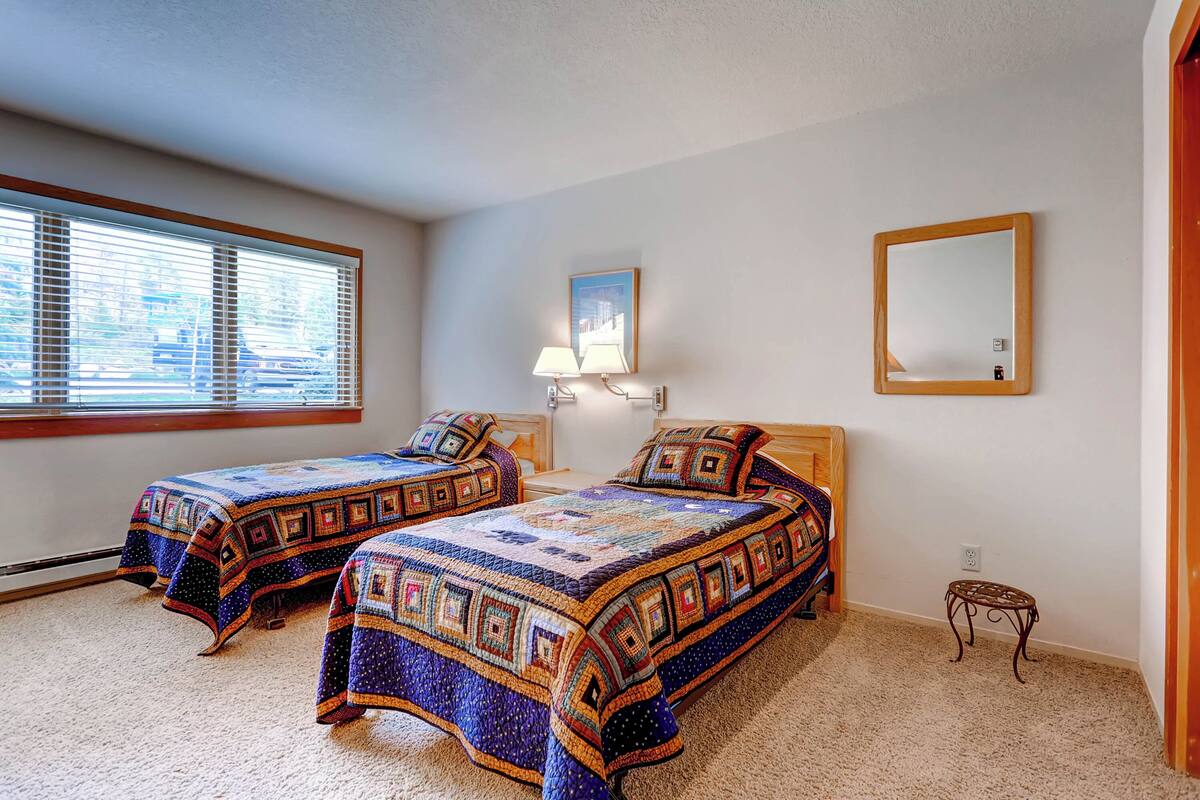 A bedroom with two beds, a nightstand, a lamp, a mirror, and a window with blinds.