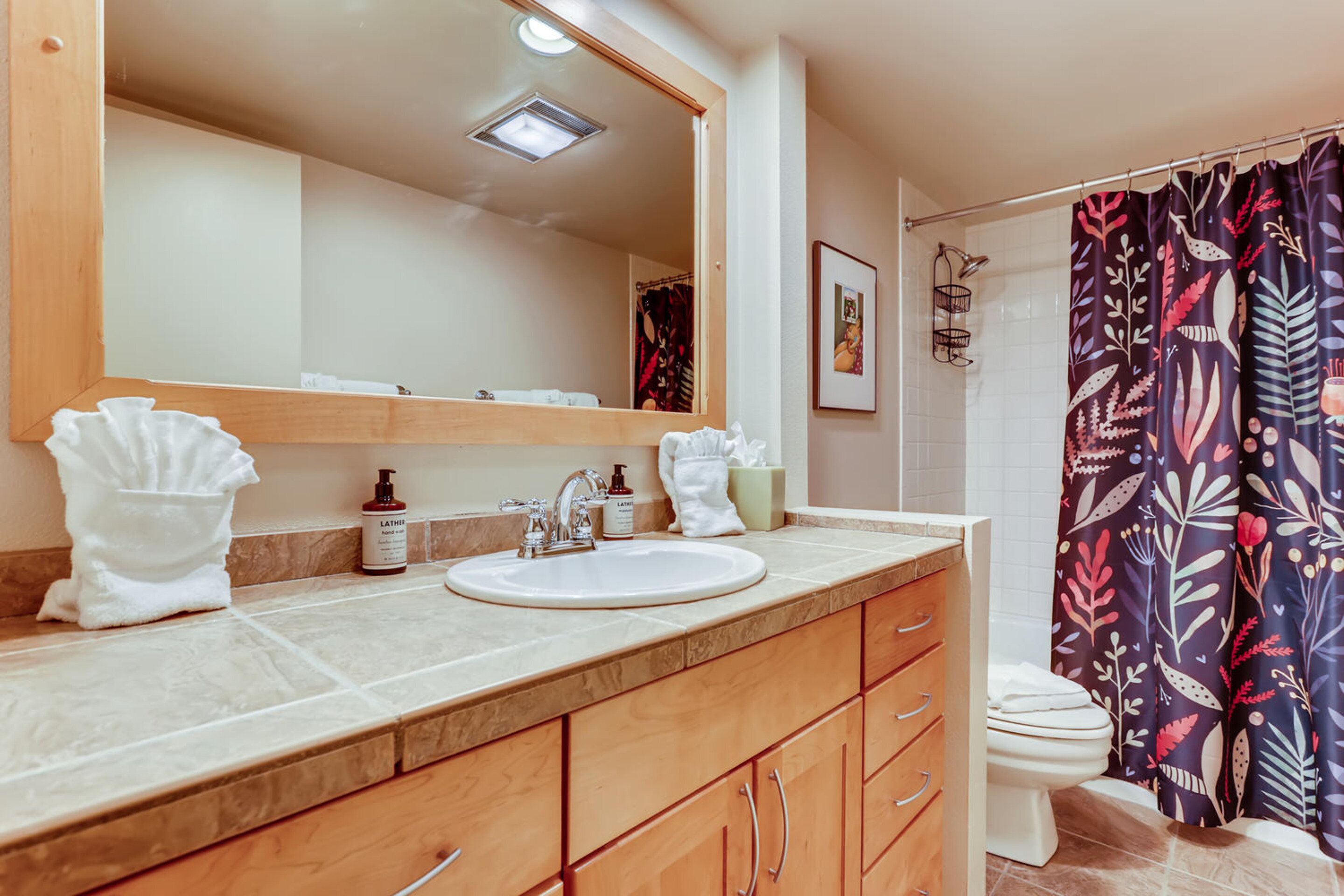 A bathroom with a sink, mirror, wooden cabinets, a toilet, and a shower curtain with a floral pattern.