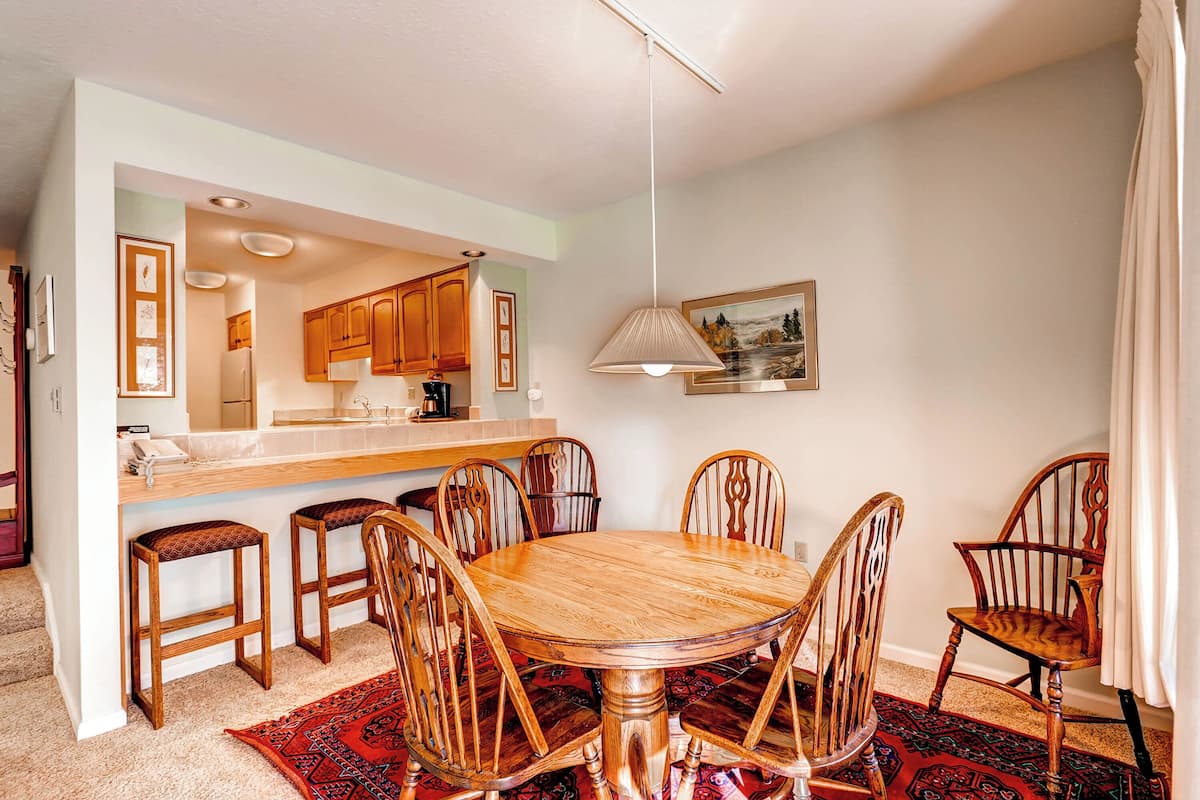 A dining area with a wooden table and chairs, a kitchen in the background, and a hanging light fixture.