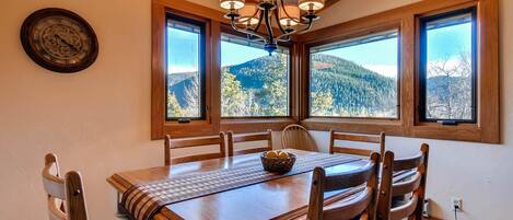 A dining area with a wooden table, chairs, a chandelier, and large windows with a mountain view.