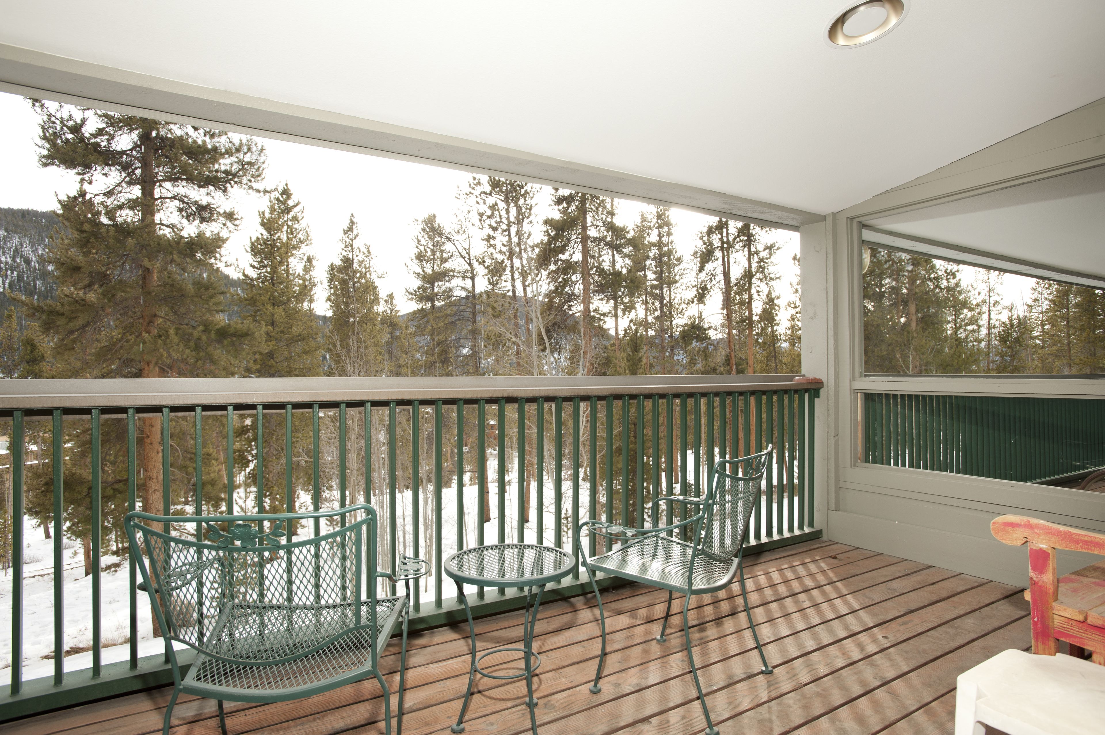 A wooden deck with metal chairs and a table, overlooking a snowy landscape with trees.