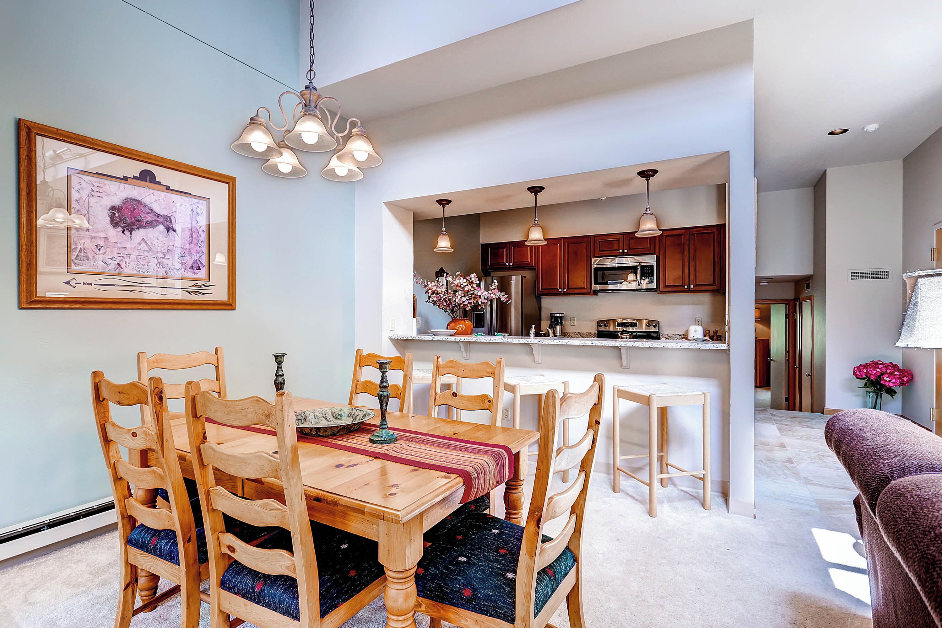 A dining area with wooden chairs and a table, a kitchen with wooden cabinets and modern appliances, and a hanging light fixture.