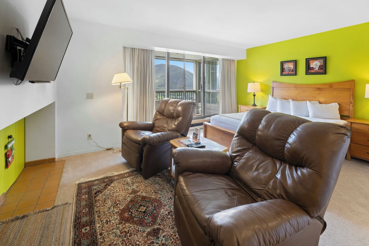 A hotel room with a bed, two leather sofas, a television, a window with curtains, and a view of a mountain.
