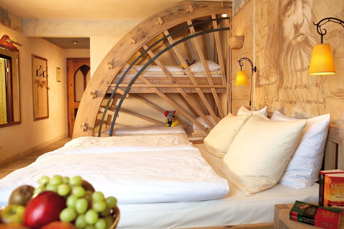 A room with a bed, a wooden arched window, and a fruit bowl on the bedside table.