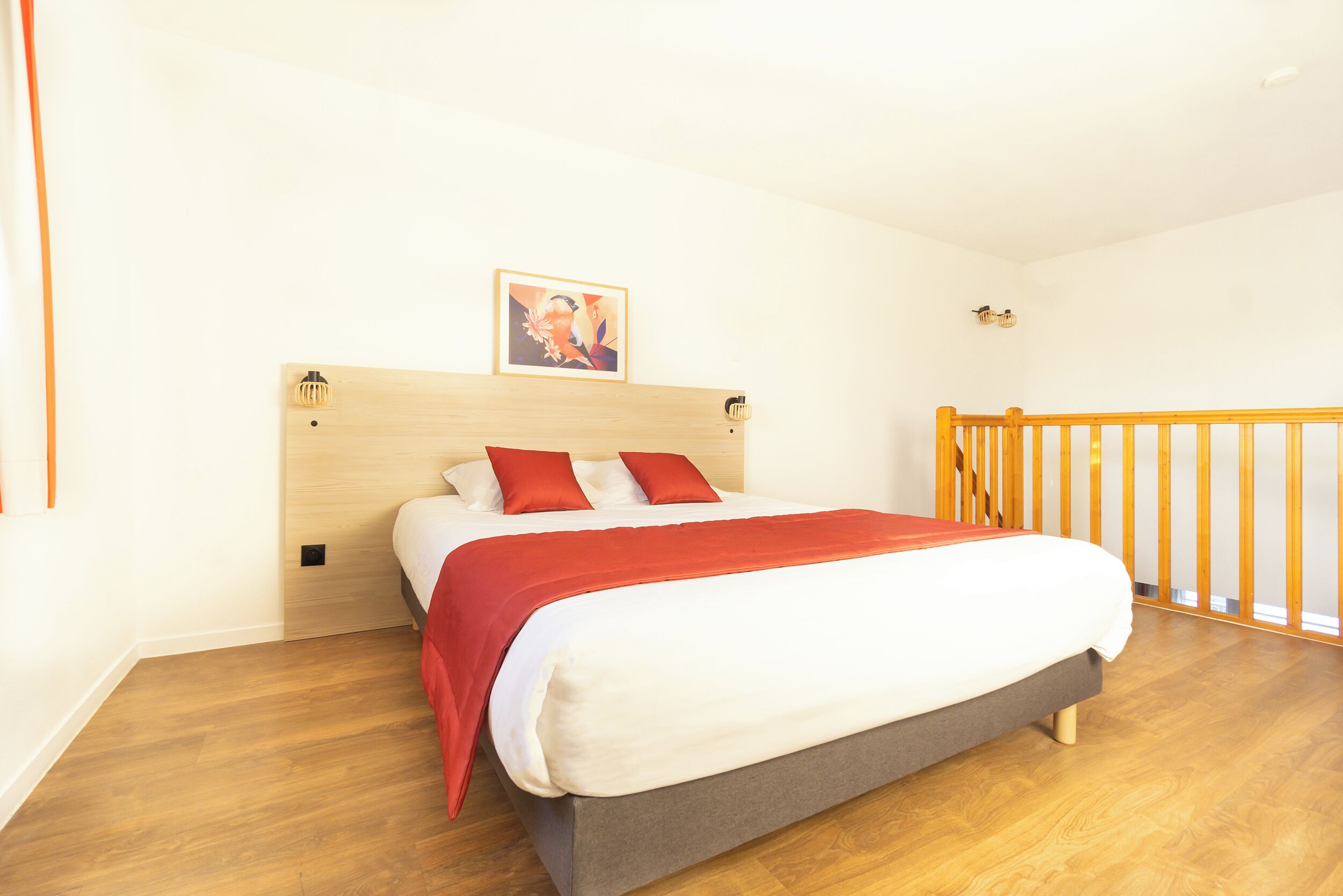 Un dormitorio con cama de madera, ropa de cama roja y blanca, cabecero de madera, un cuadro en la pared y barandilla de madera.