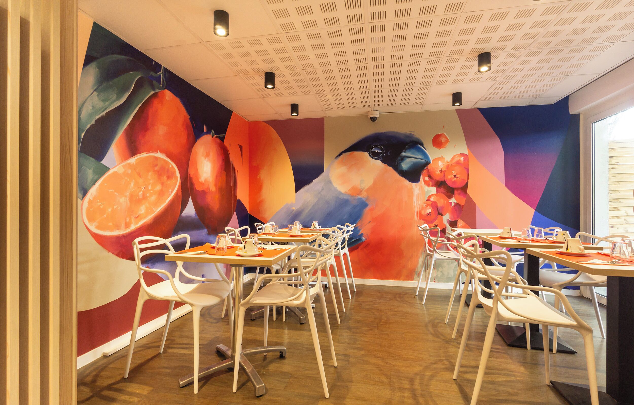 A dining area with tables and chairs, a ceiling with recessed lighting, and a mural with fruit and a bird.