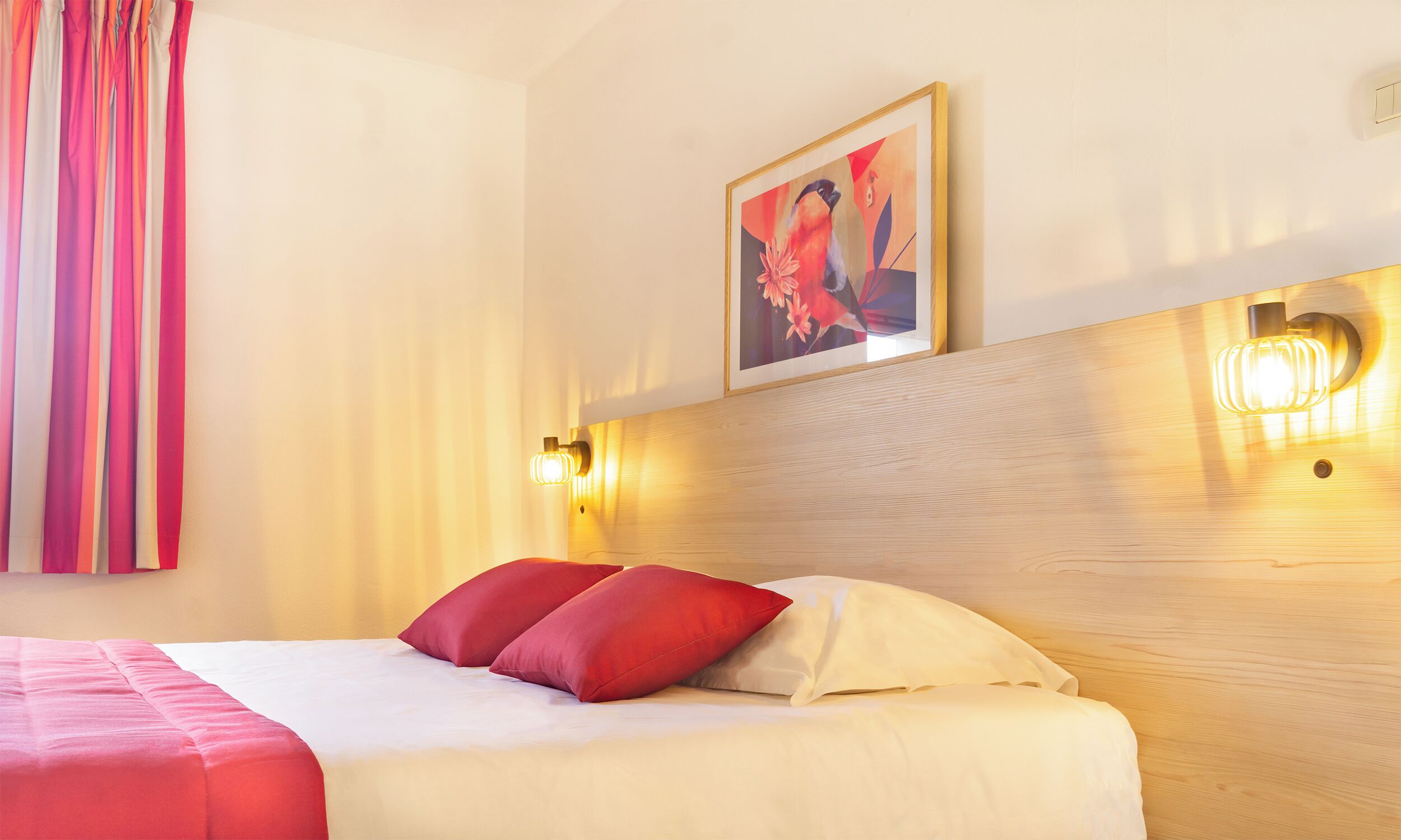 Habitación de hotel con cama, ropa de cama roja y blanca, un cuadro en la pared y cabecera de madera.
