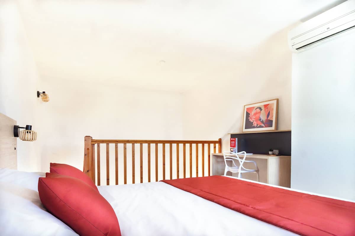 A bedroom with a wooden headboard, a bed with red and white bedding, a desk with a chair, and a framed picture on the wall.