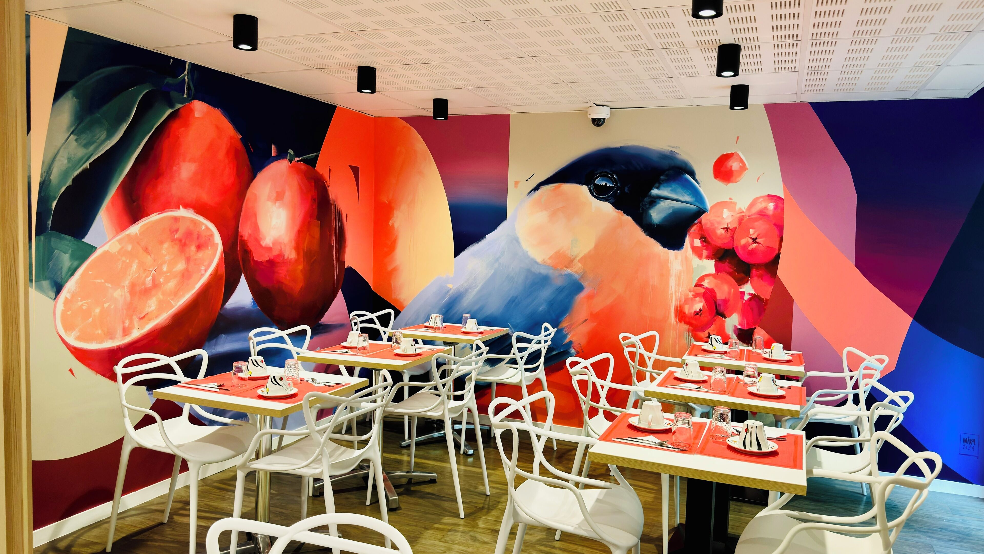 Ein Restaurant mit weißen Stühlen und Tischen, einer Decke mit eingelassener Beleuchtung und einem farbenfrohen Wandgemälde.