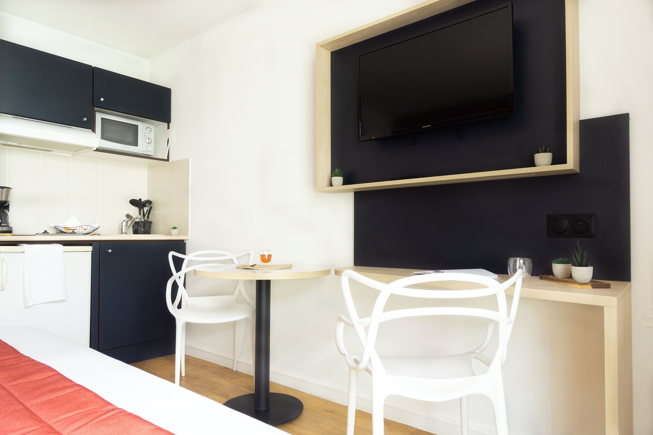 A modern kitchen with a microwave, a TV mounted on the wall, two white chairs around a small table, and a red rug on the floor.